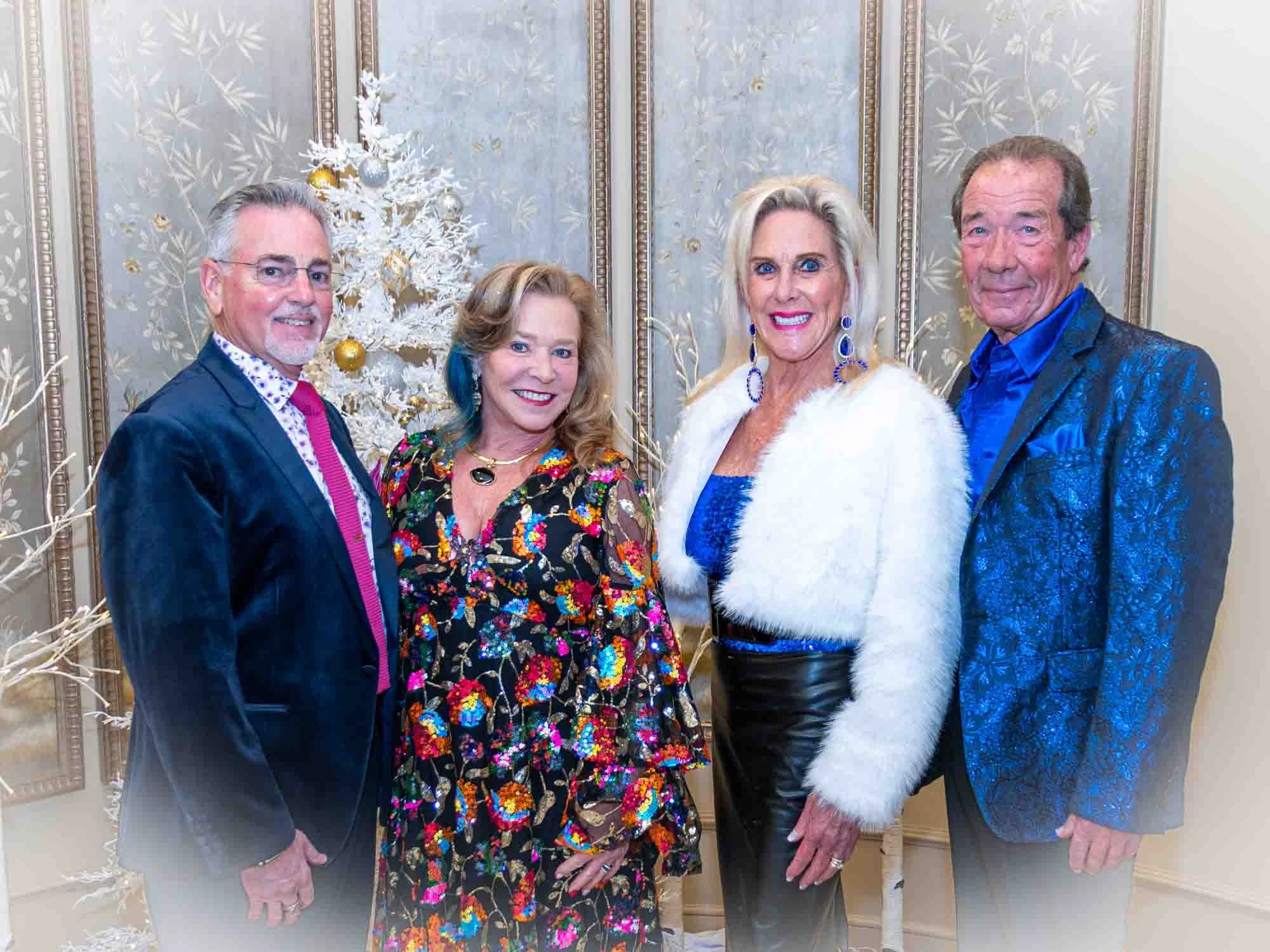 Four people, two men and two women, posing in front of a decorated white Christmas tree, wearing colorful dressy outfits. The background features ornate wallpaper.