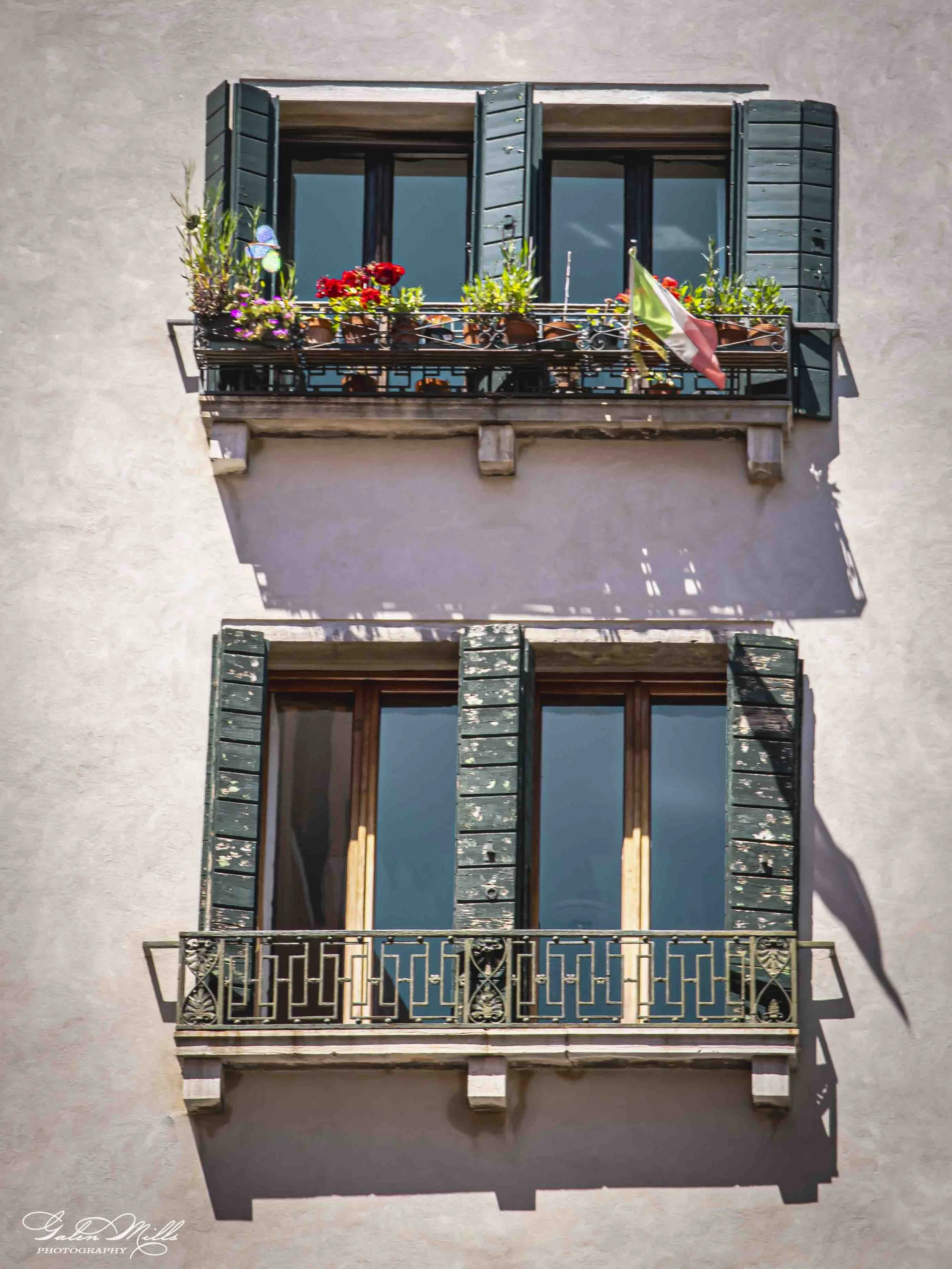 A photo of two small balconies on the exterior of an apartment building, with open green shutters, flower pots, and a small Italian flag, with shadows on the wall.