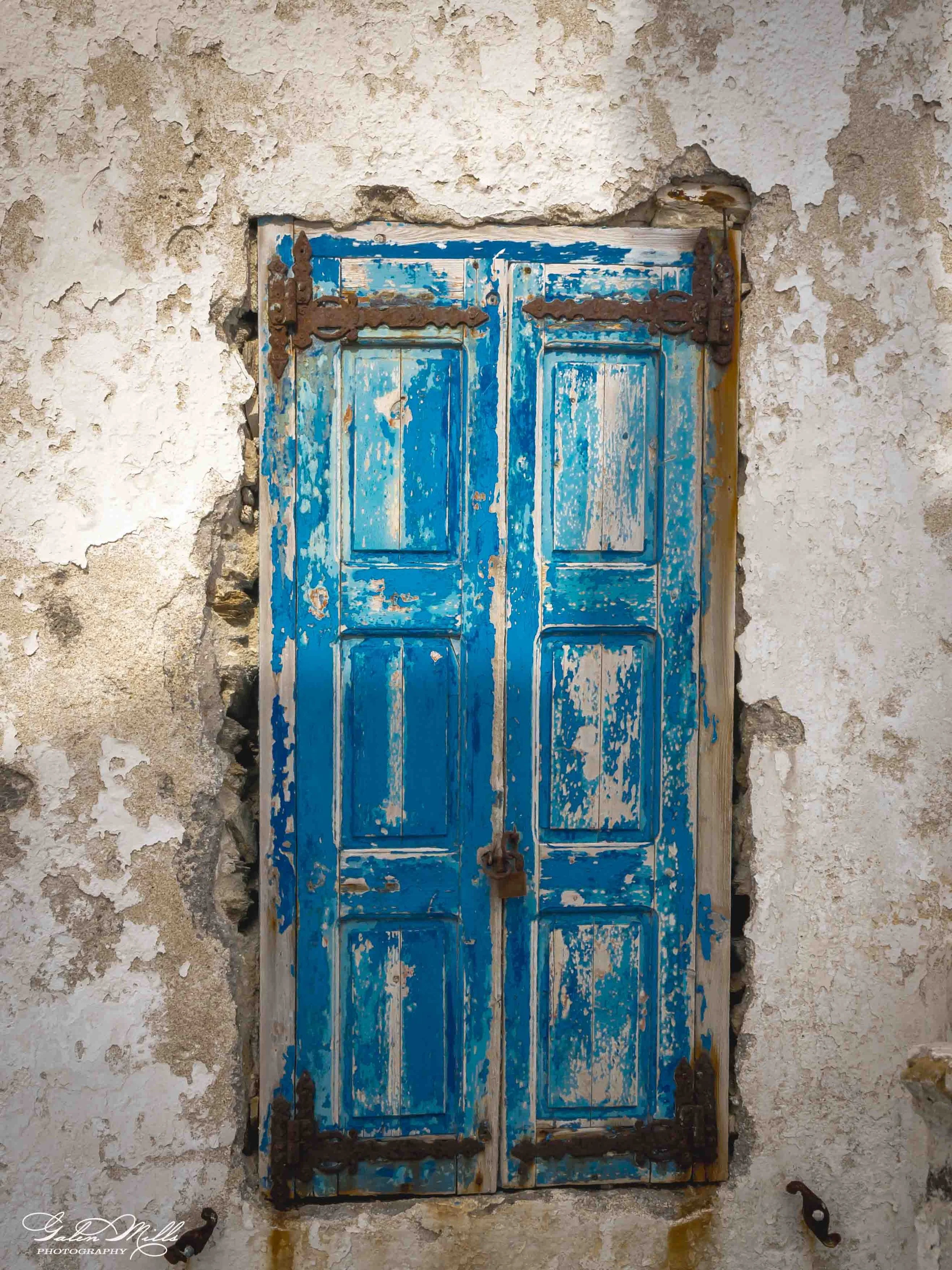 An old wooden door with peeling blue paint set in a weathered wall with peeling white plaster.