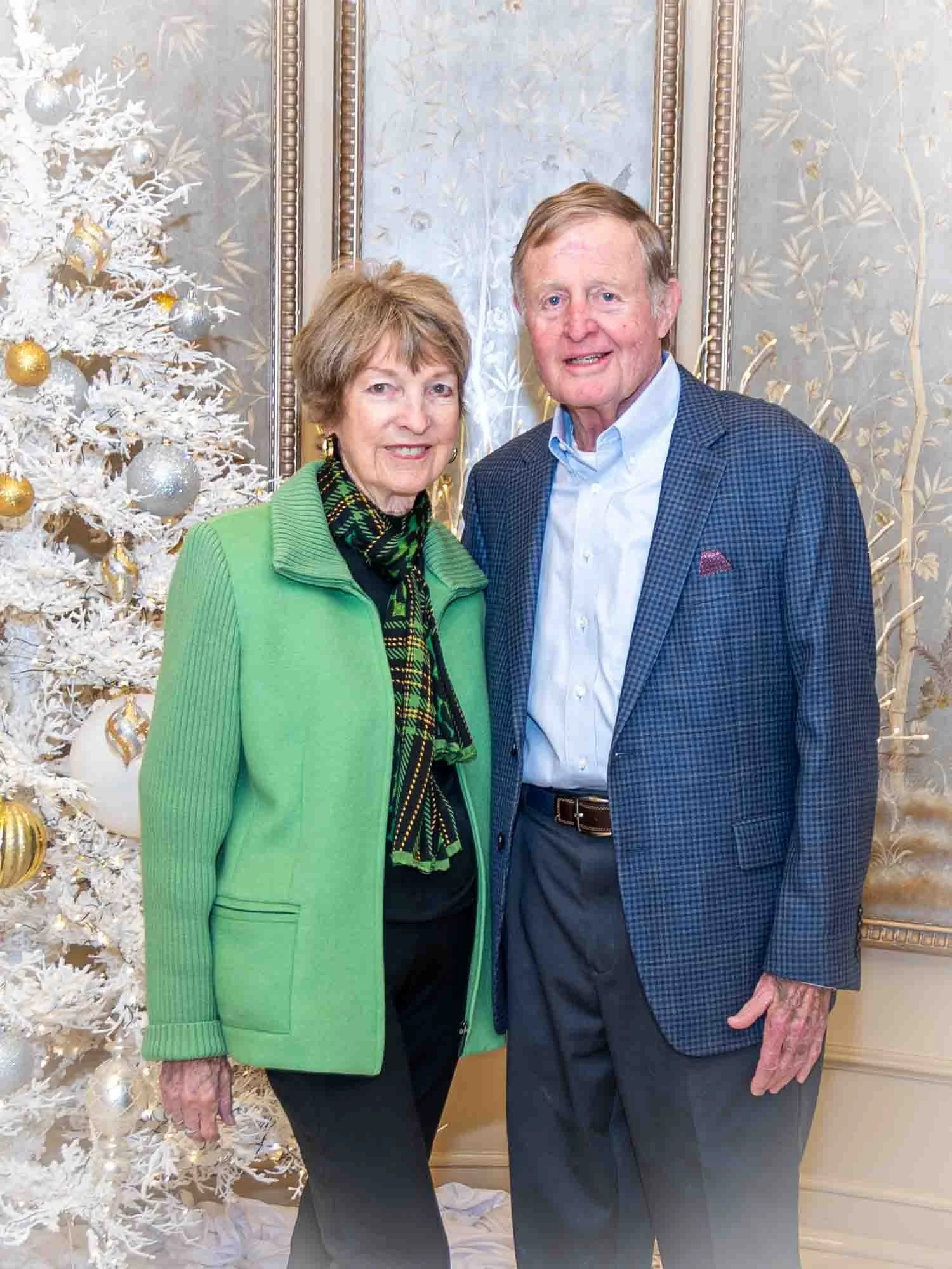 An elderly couple standing in front of a decorated white Christmas tree, with silver and gold ornaments, in an elegant room with ornate wallpaper.