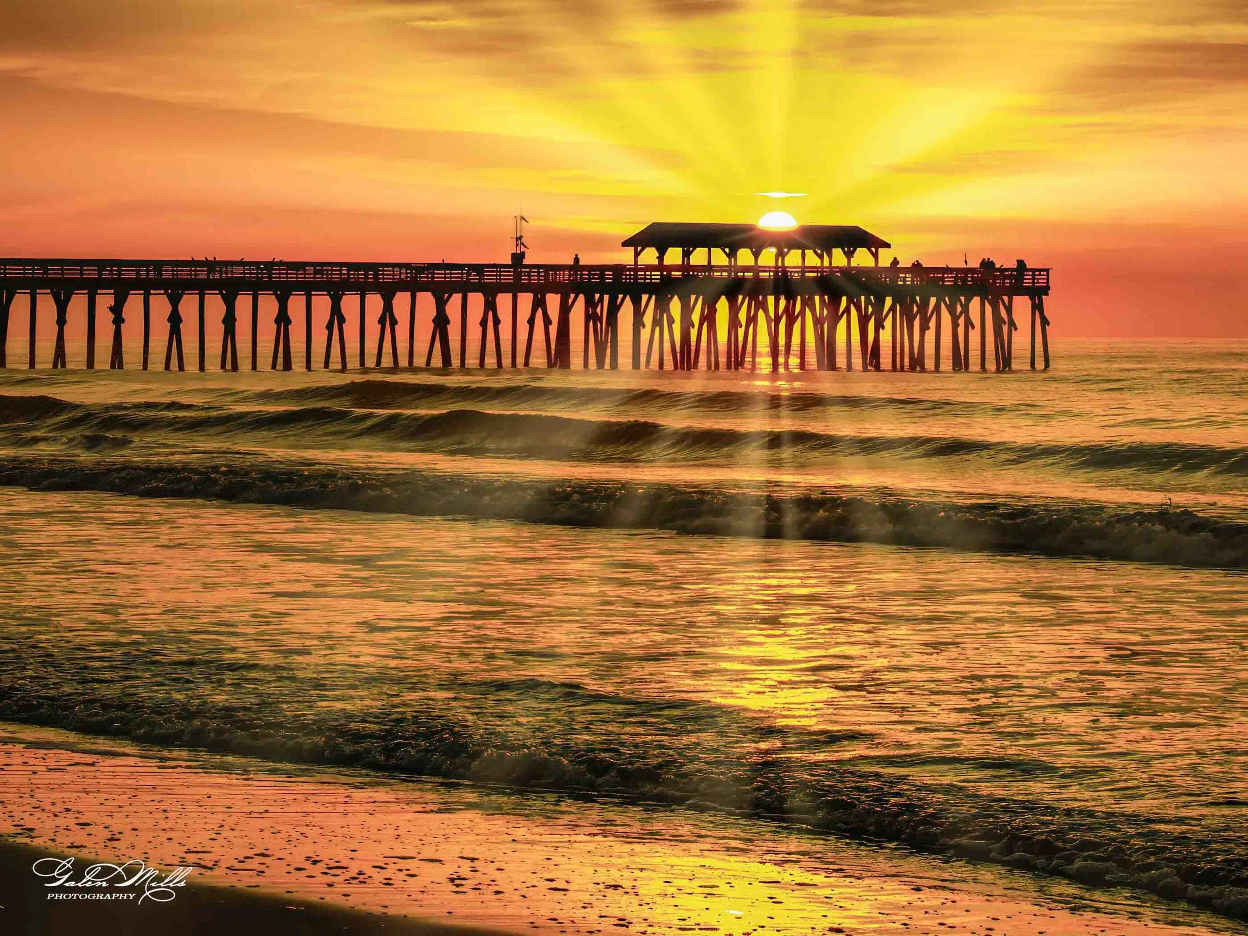 A pier extending into the ocean at sunset with the sun setting behind a shelter on the pier, casting rays and reflections on the water.