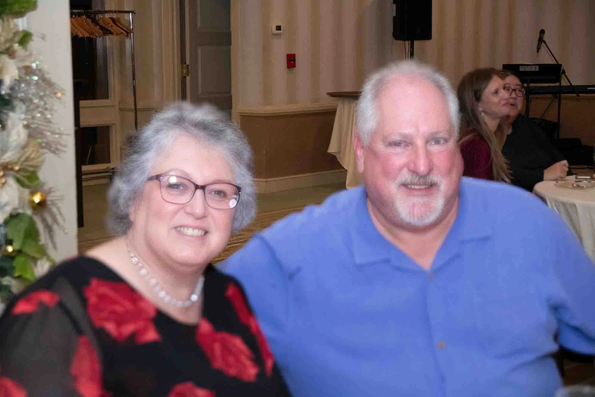 Smiling older couple at a formal event, woman wearing glasses and a black dress with red floral print, man in a blue shirt, indoor setting with tables in the background.
