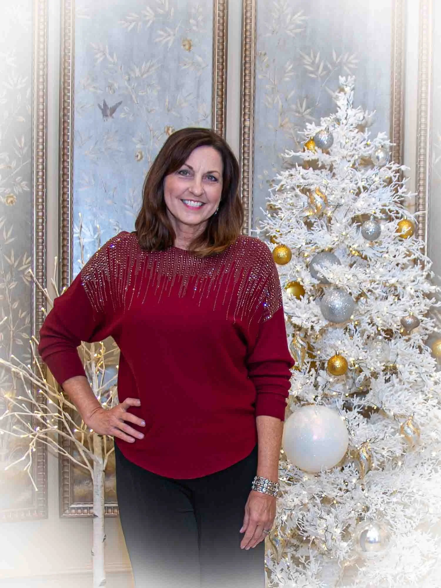 Smiling woman in a red sweater standing next to a white Christmas tree with gold and silver ornaments.