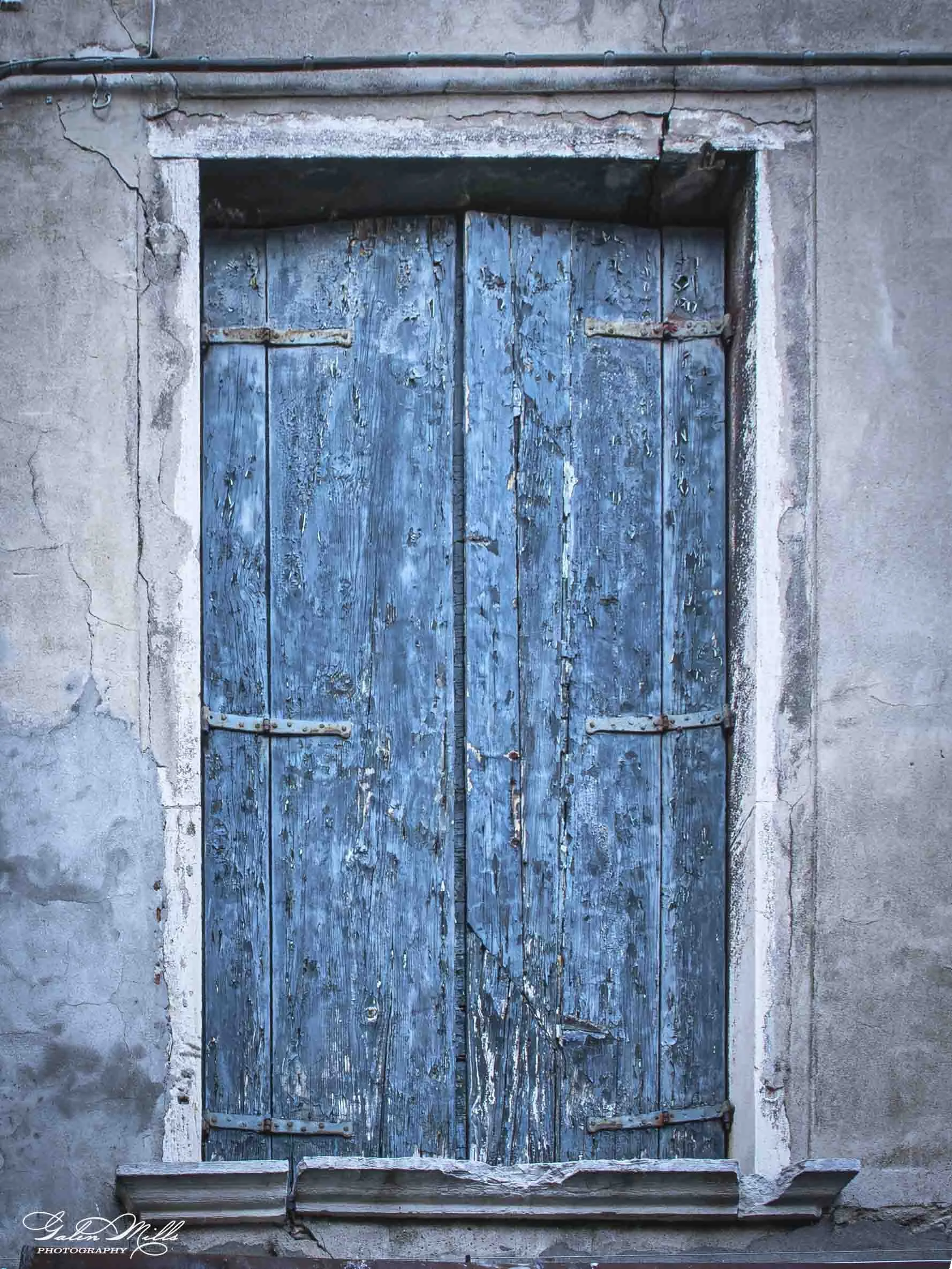 An old blue wooden door with peeling paint, set in a cracked concrete wall, with metal hinges and a metal bar crossbar across the middle.