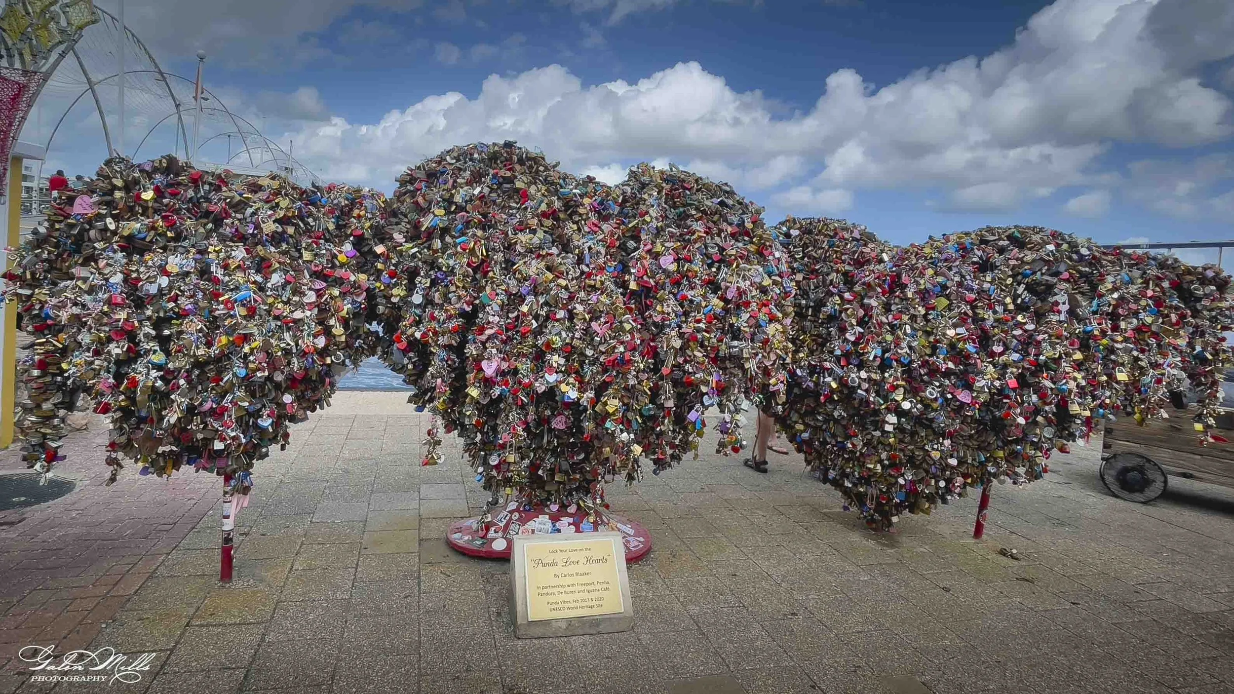 A sculpture of a bear made entirely of colorful locks and padlocks, located outdoors near a beach under a partly cloudy sky. There is a sign at the base of the sculpture that reads 'Panda Love Hearts' by Carlos Beukelaer, with additional details abou