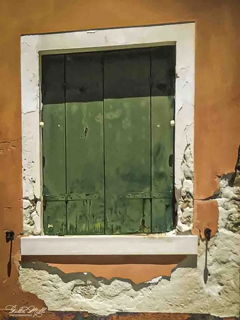 A small window with green shutters, set in a rustic wall framed by white stone and orange plaster.