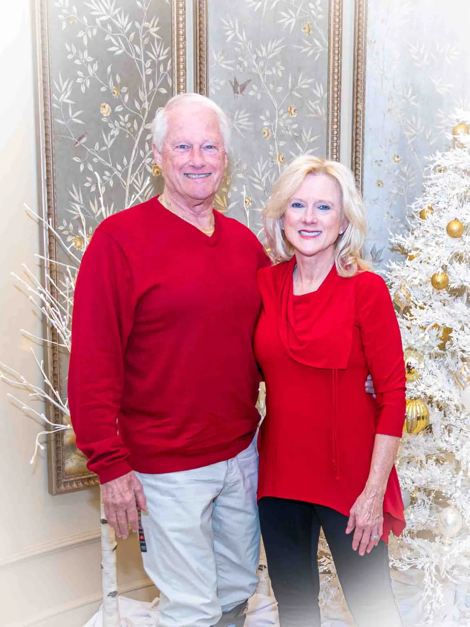 An older couple dressed in red holiday attire, standing in front of a white Christmas tree with gold ornaments, next to an ornate wall with floral patterns.