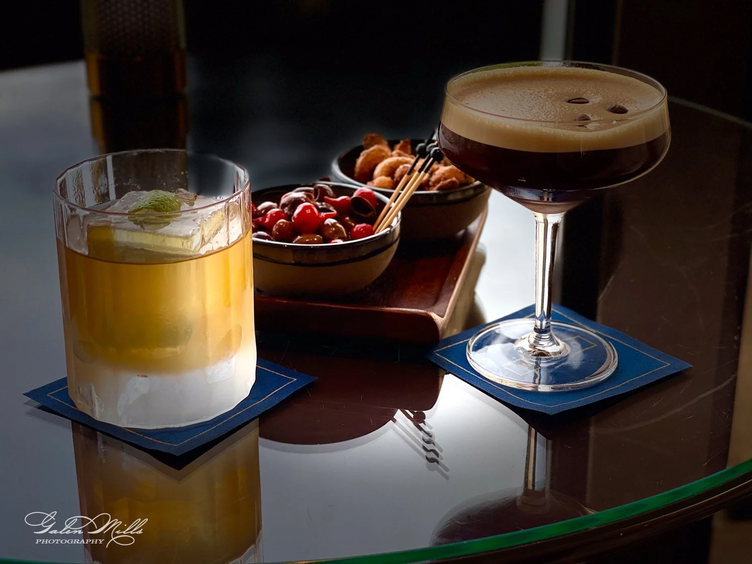 Two cocktails on a table with nuts and cherries.