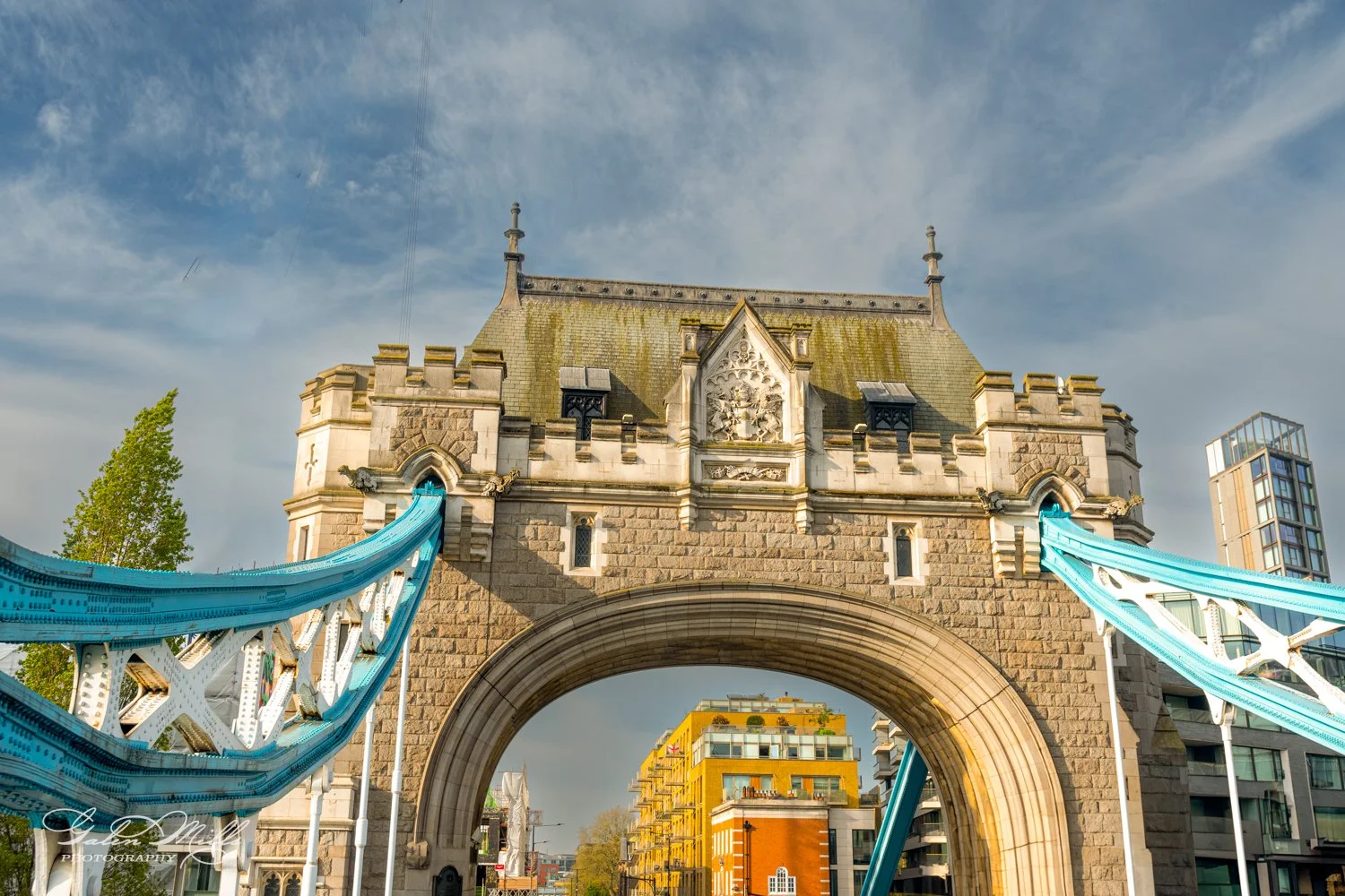 Close-up of the central arch of Tower Bridge in London, showing decorative stonework, blue suspension cables, and surrounding modern buildings against a cloudy sky.