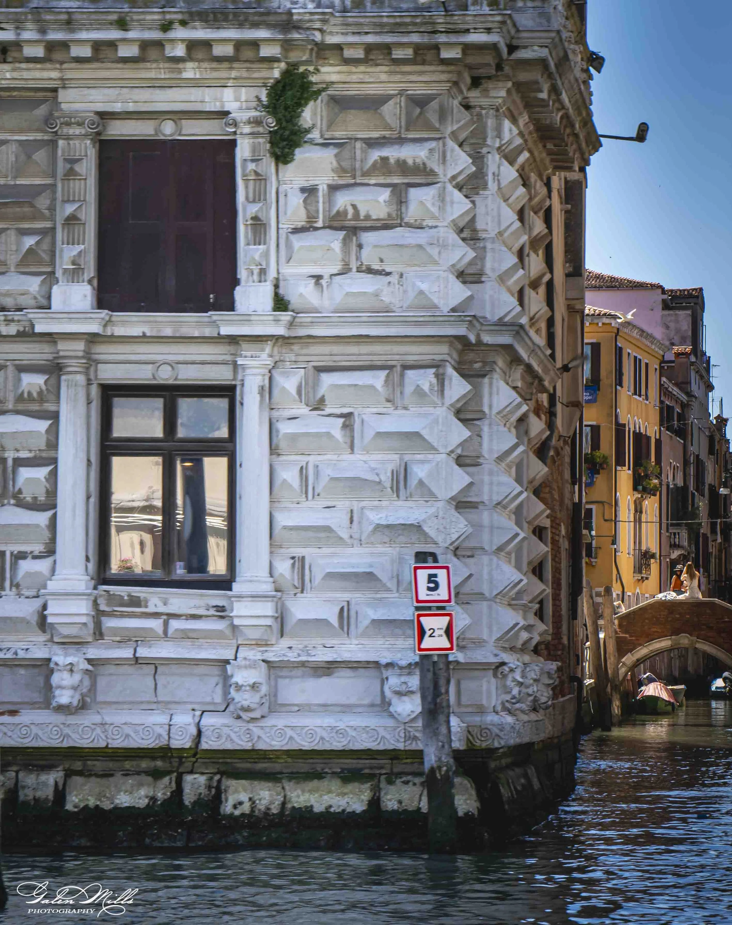 The front of an ornate, historic building with marble-like decorative stonework, located alongside a canal. Buildings are visible in the background, and a small boat is partially seen on the water below. There are signs on the building, including a s