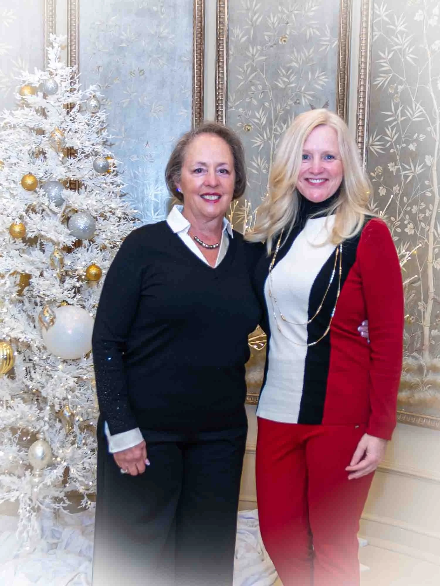 Two women standing in front of a decorated Christmas tree, one wearing a black and white outfit, the other in a red and black ensemble, set in an ornate room with patterned wallpaper.