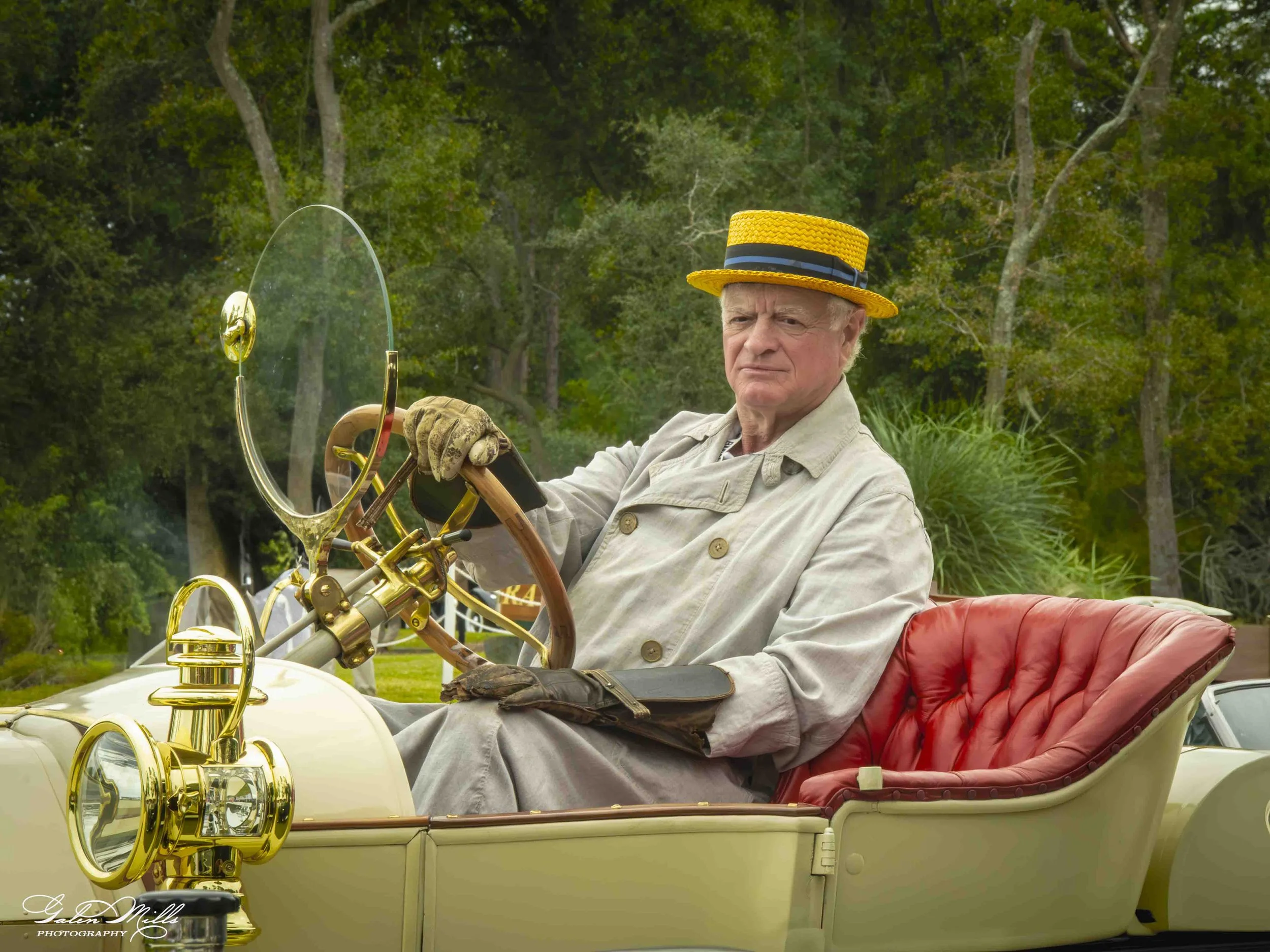 An elderly man wearing a yellow straw hat with a black ribbon, beige jacket, and leather gloves, sitting in a vintage fire truck with a red leather seat, holding the steering wheel with a serious expression, surrounded by green trees.