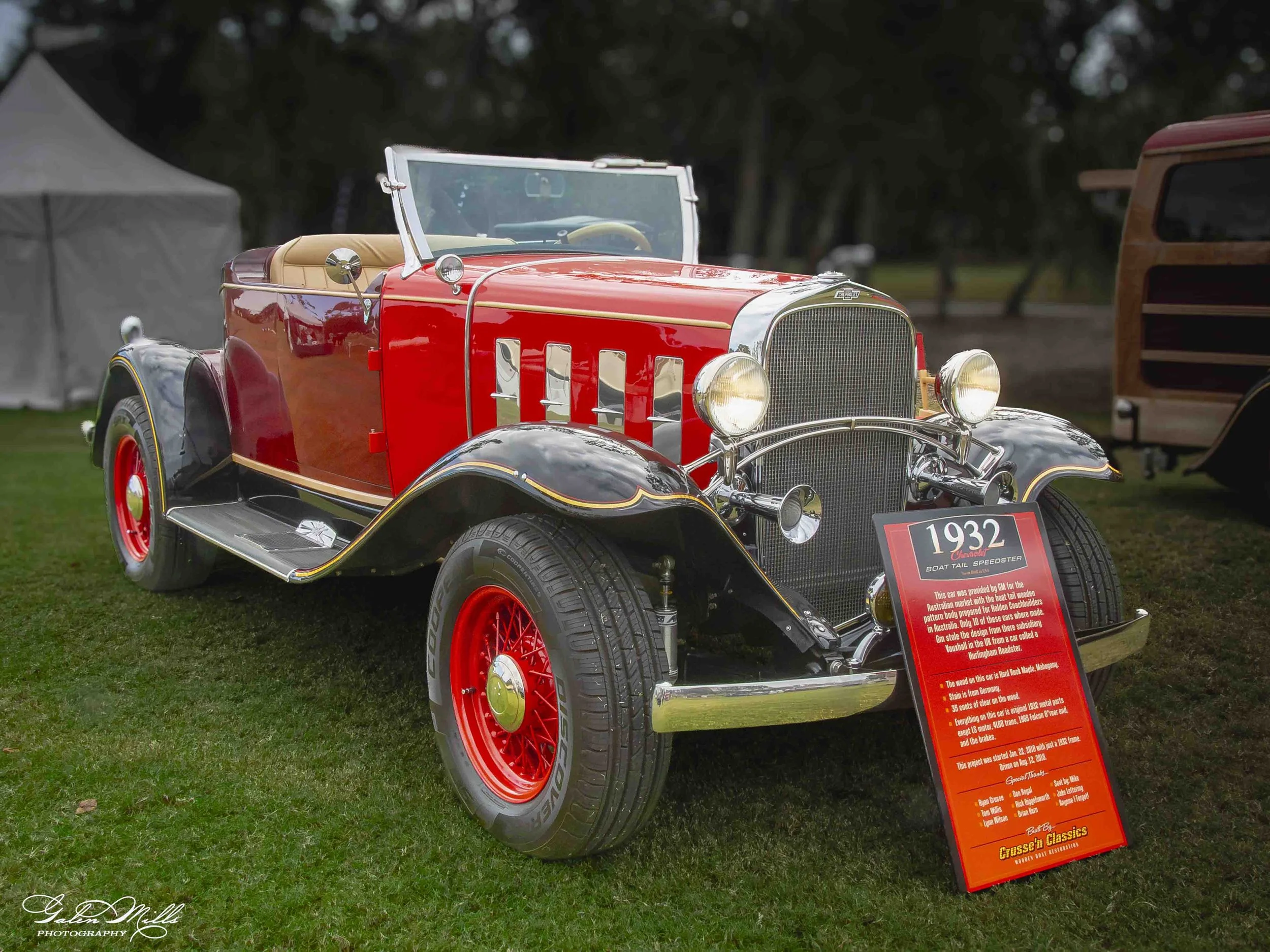 A vintage red and black 1932 Chevrolet boat tail speedster car on display at an outdoor event with a sign providing details about the vehicle.