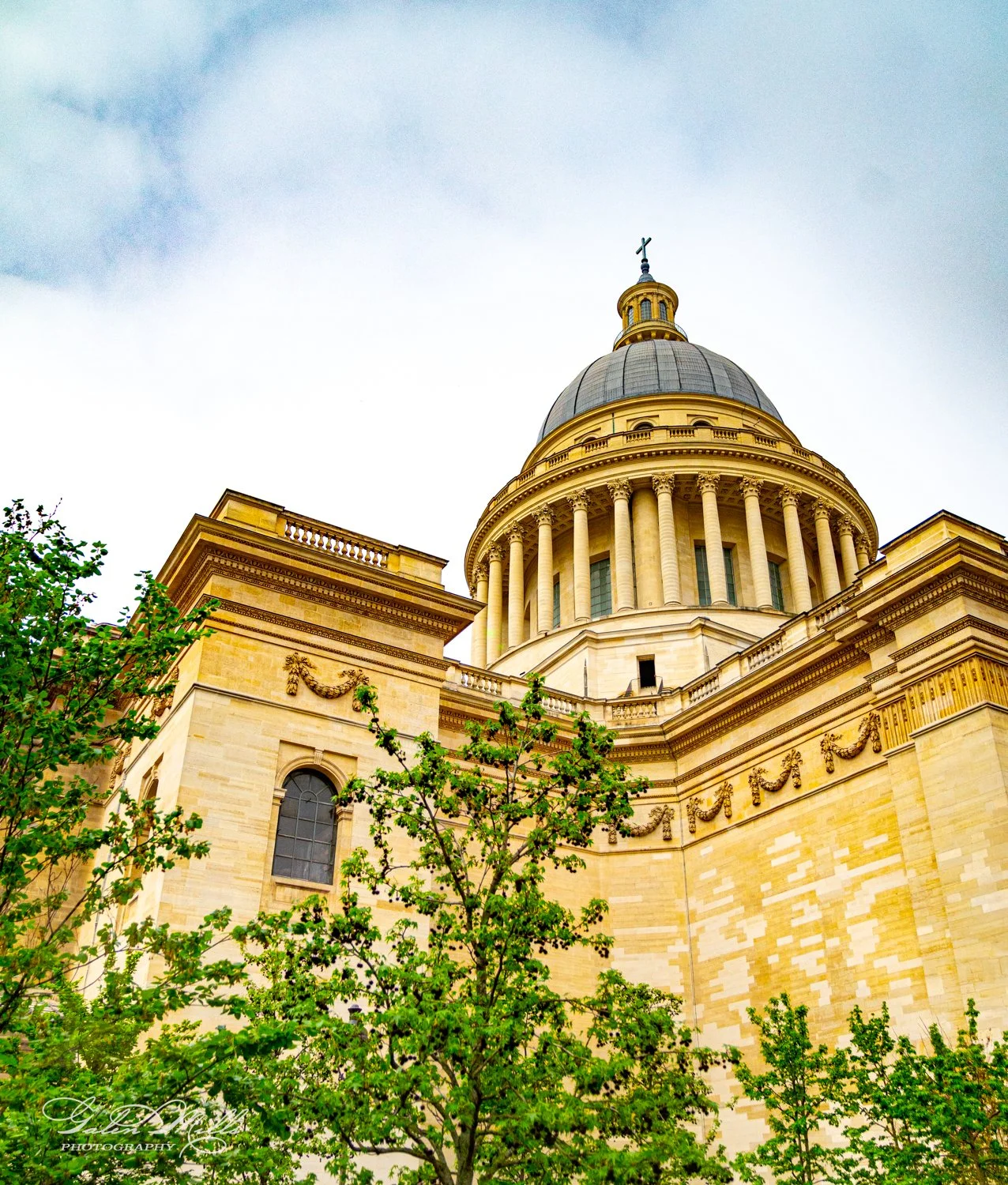 A grand historical building with a large dome and classic architectural features, including columns, surrounded by green trees, under a partly cloudy sky.