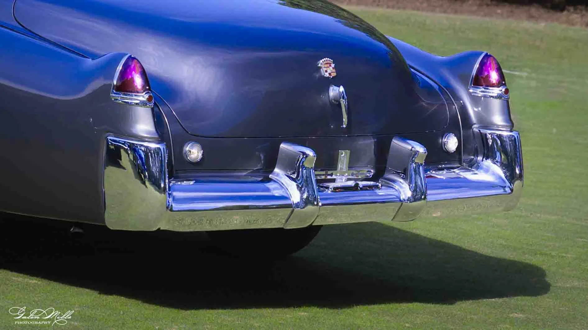 Close-up of the rear end of a vintage black Cadillac car with chrome bumper, taillights, and Cadillac emblem, parked on grass.