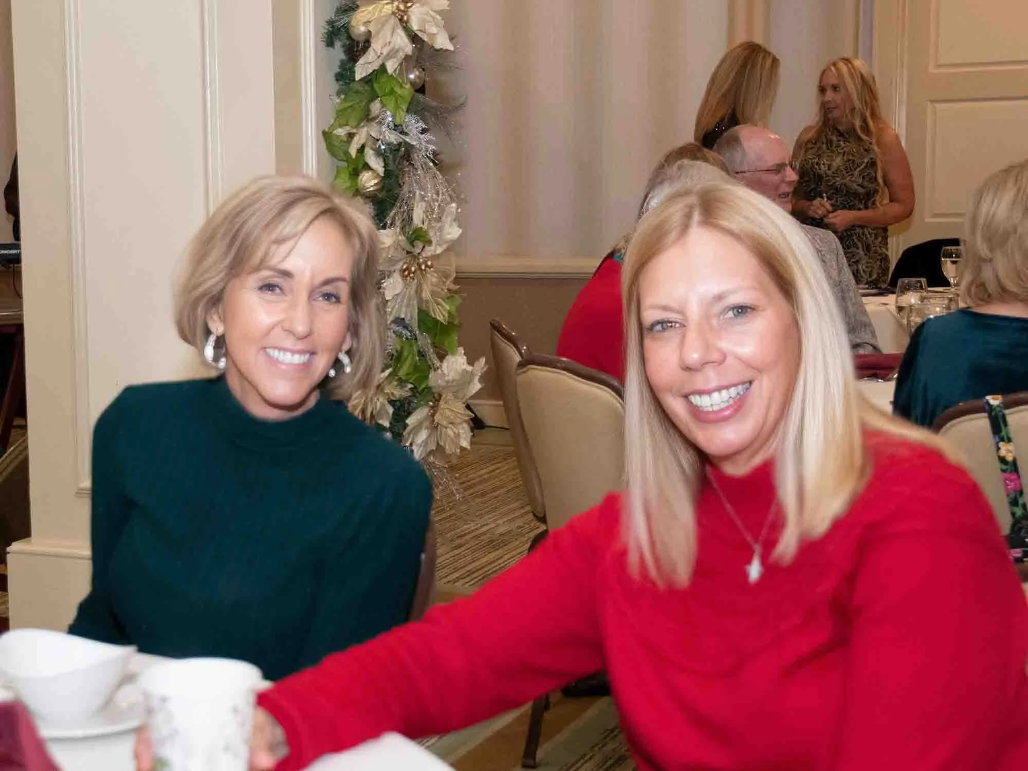 Two women smiling at a holiday event, one in a green top and the other in a red top, sitting at a table with cups and bowls. A decorated Christmas garland is visible in the background.