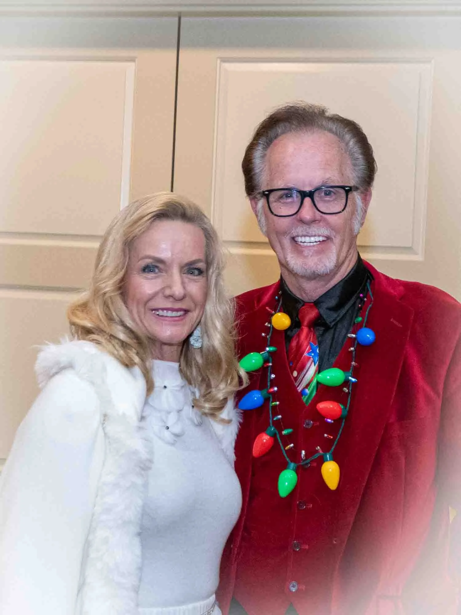 A man and a woman in festive attire, with the man wearing a red suit and a necklace of colorful lights.