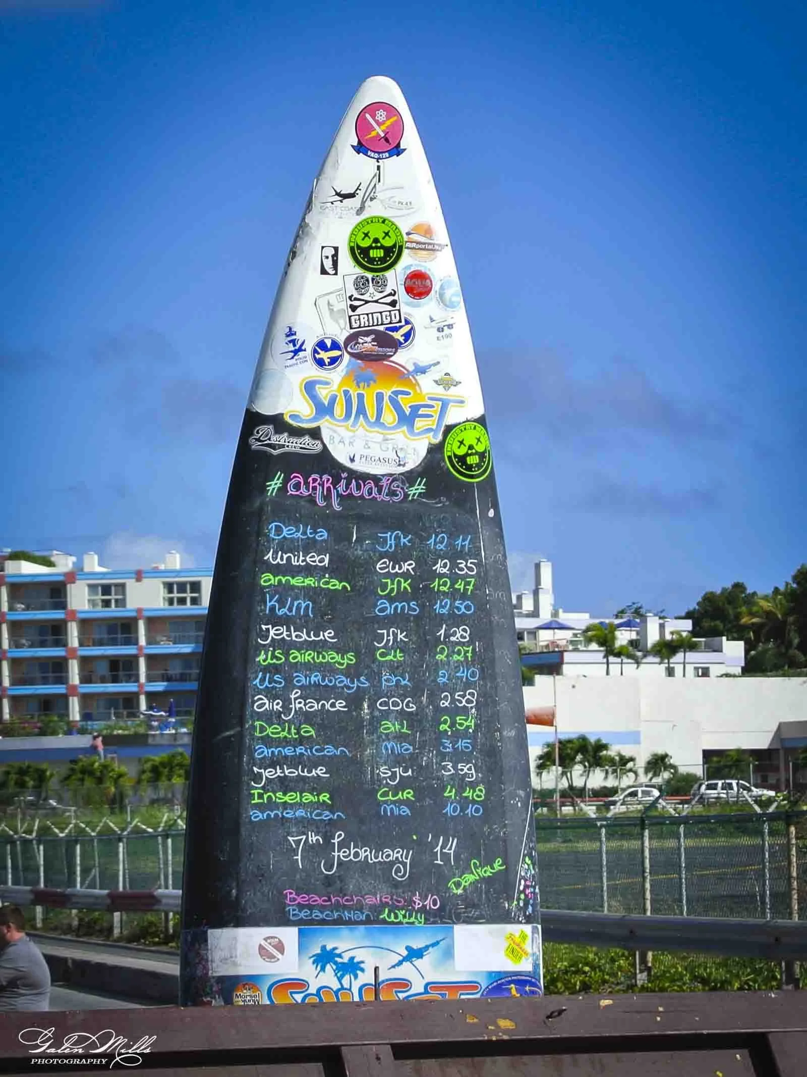 A large blackboard menu with colorful handwritten pricing details for various airlines and destinations, decorated with airline stickers and mounted on a pole outdoors, with buildings and trees in the background on a sunny day.