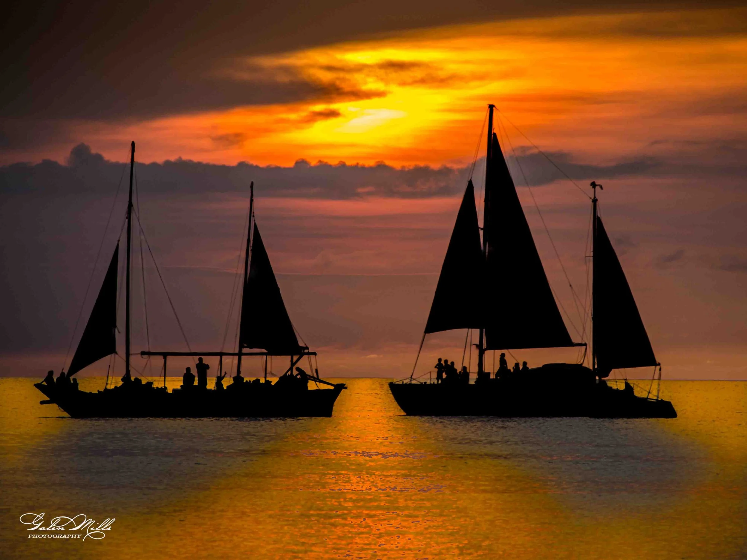 Two sailboats silhouetted against a sunset over the ocean, with people onboard and clouds in the sky.