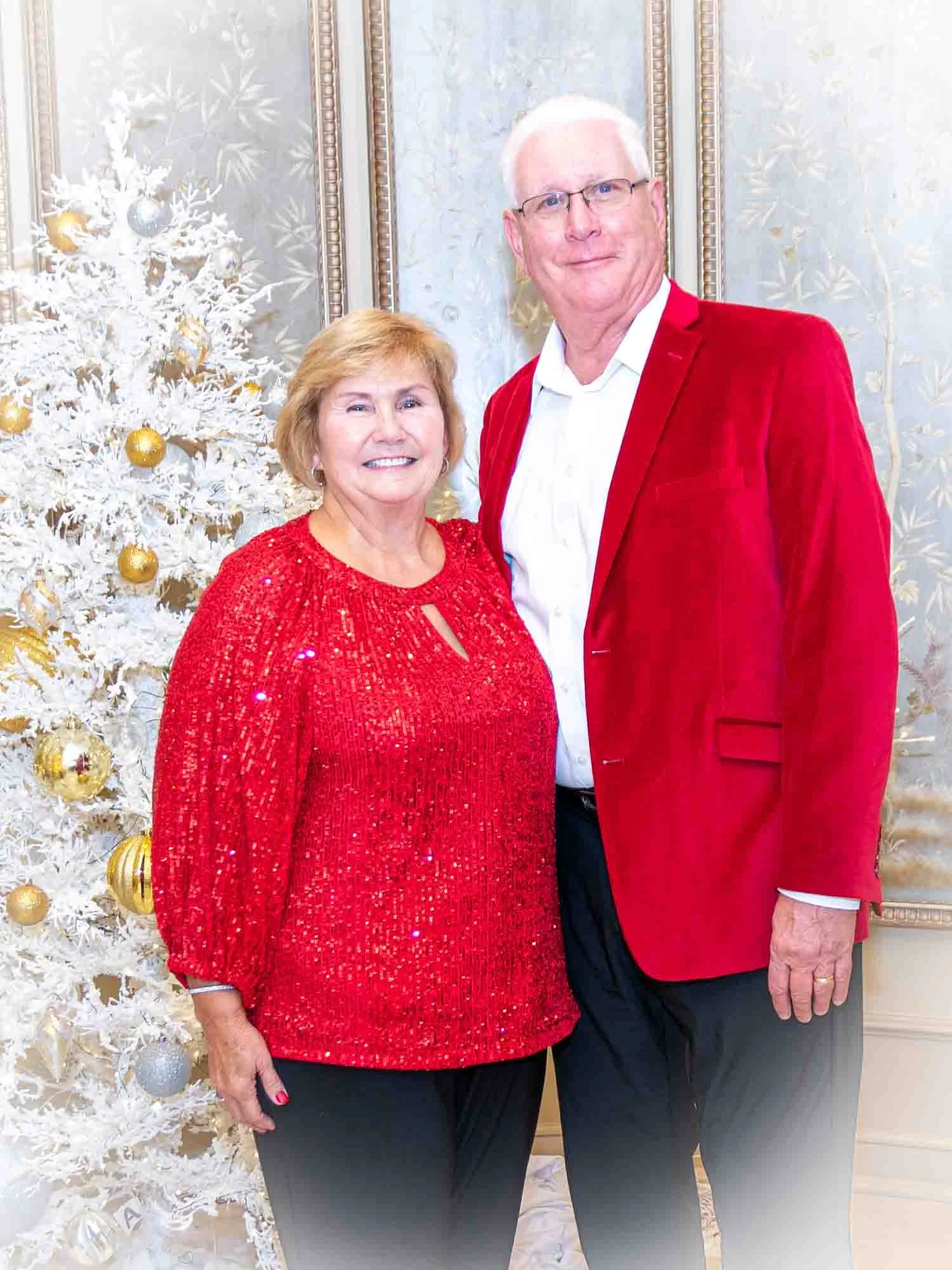 A man and a woman standing in front of a white Christmas tree with gold and silver ornaments; both are wearing red clothing.