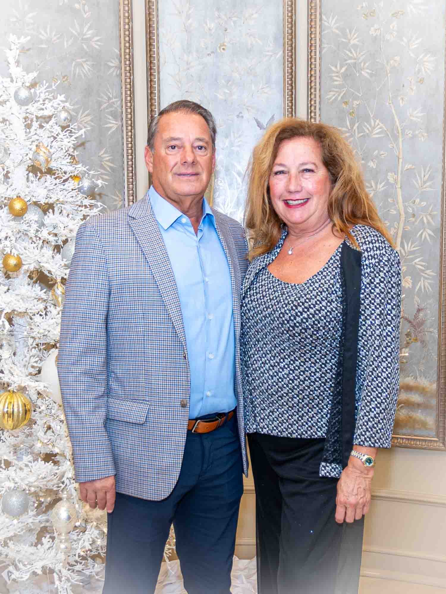 A man and woman standing together, smiling, in front of a decorated white Christmas tree. The man is wearing a checked blazer and blue shirt, while the woman is in a patterned blouse. They are indoors with elegant wallpaper in the background.