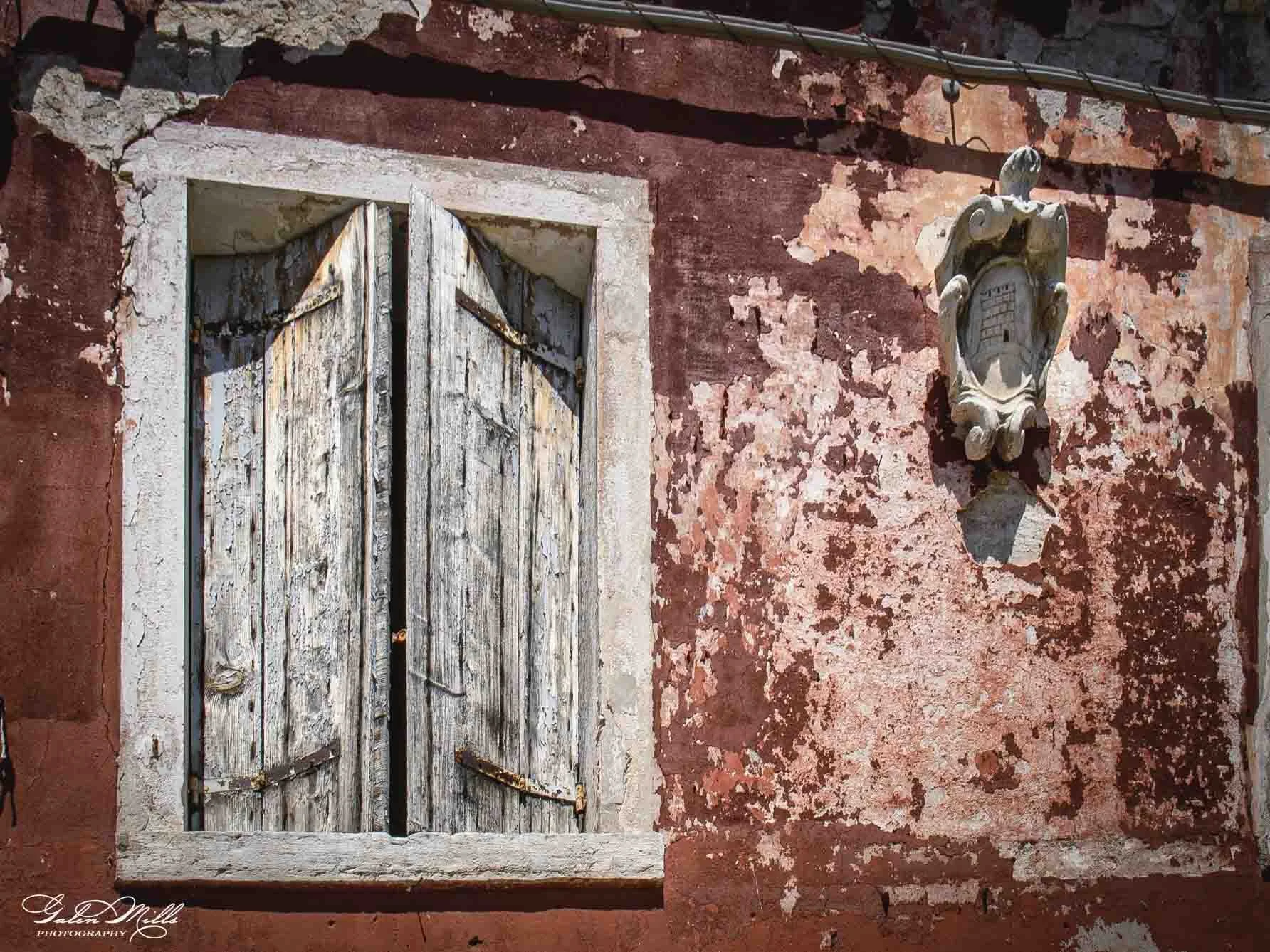 A weathered wooden window with shutters set in a red, peeling plaster wall, and an ornate stone or plaster decorative element above it.