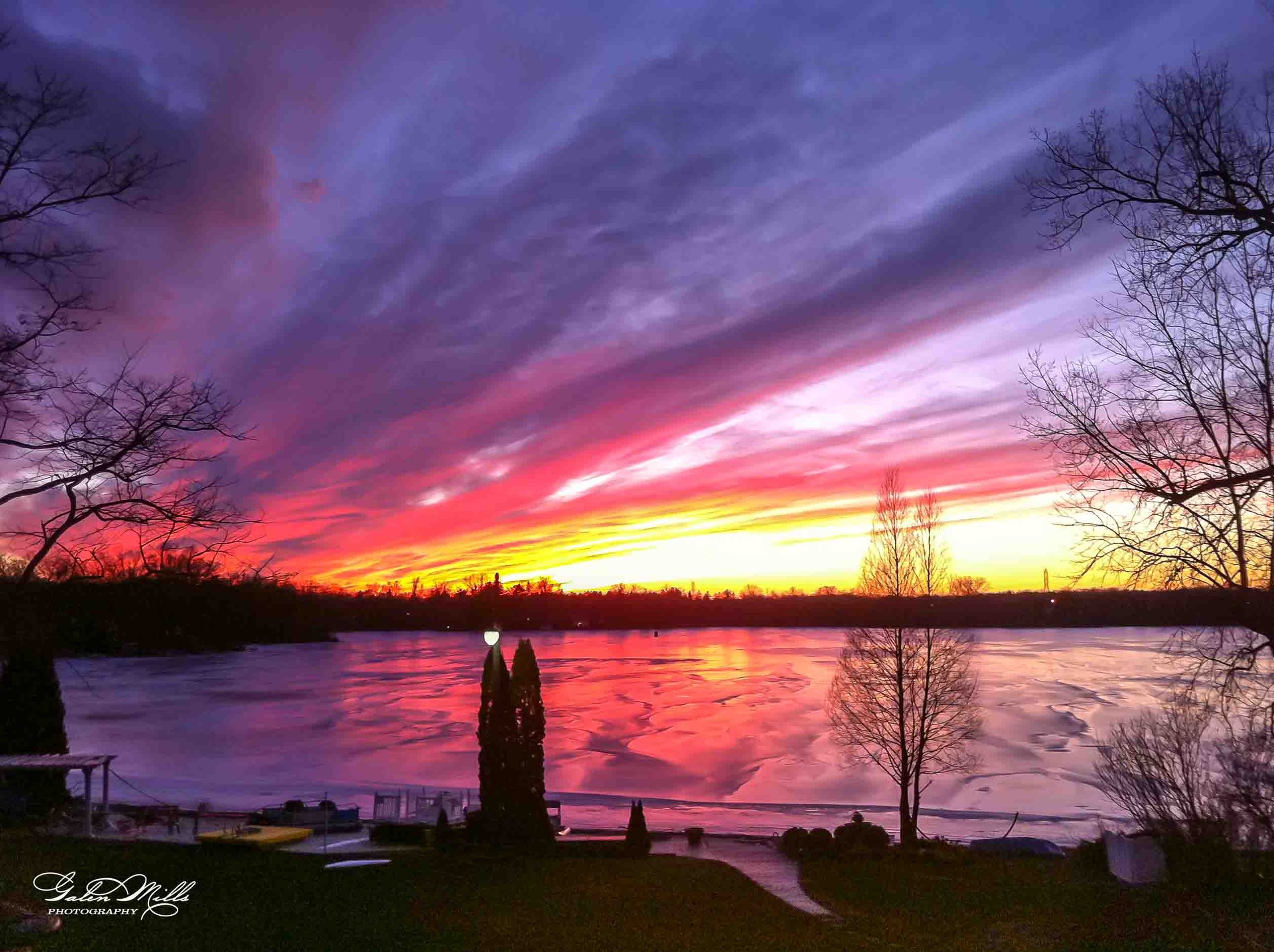 Colorful sunset over a partially frozen lake with leafless trees and shrubs, reflecting pink, purple, and yellow hues in the sky and water.