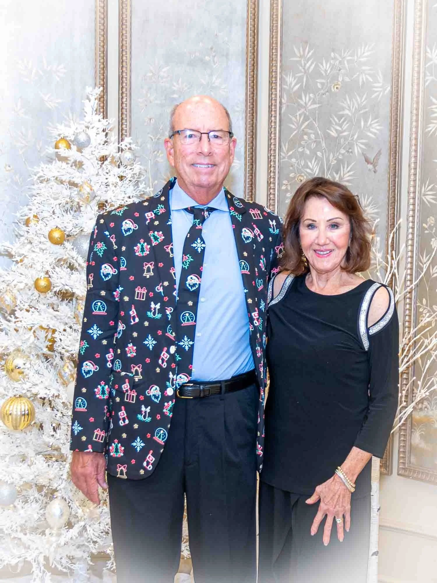 A man and woman posing in front of a white Christmas tree. The man is wearing a festive suit jacket with holiday-themed designs, and the woman is wearing a black dress.