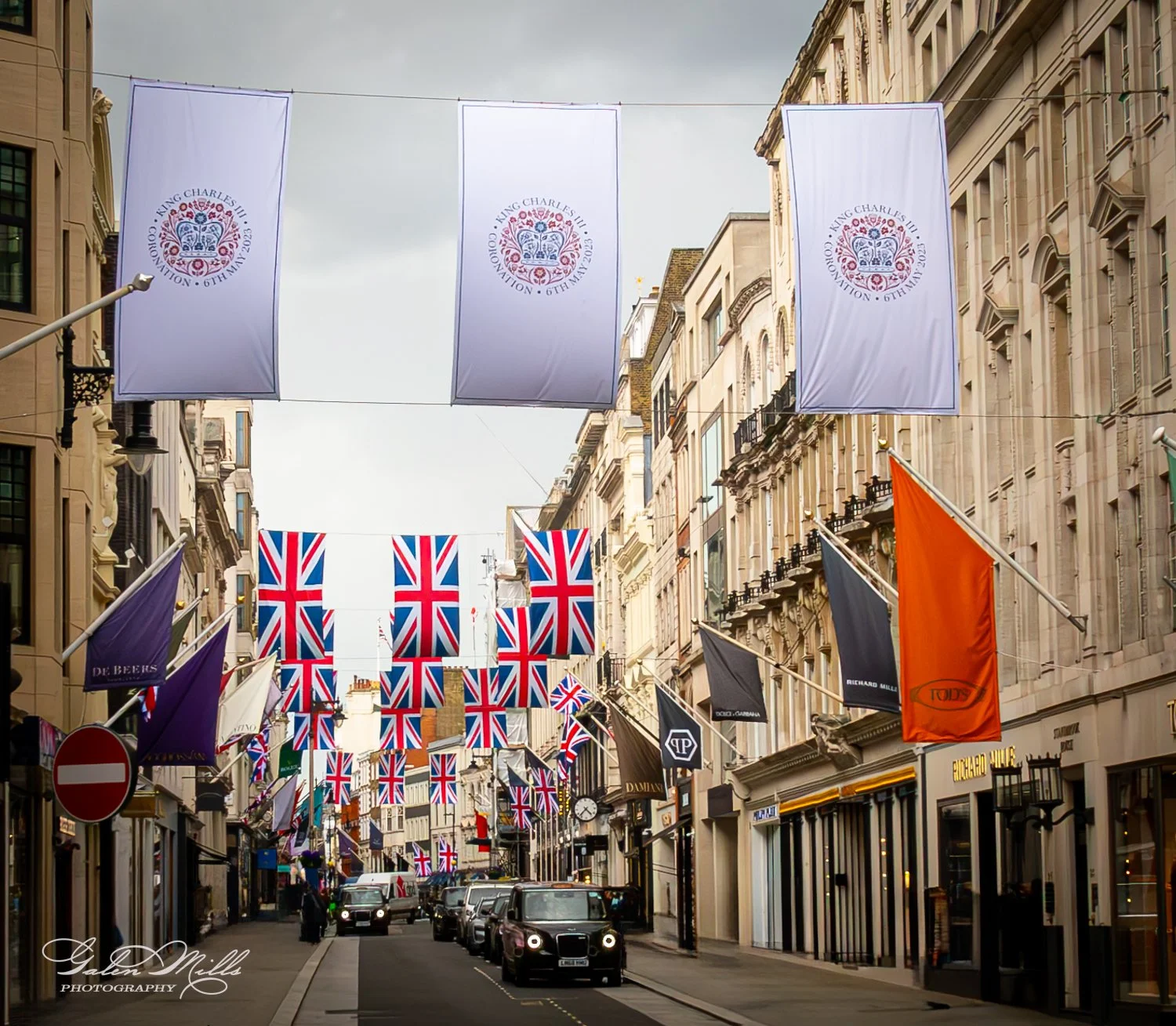 Street decorated with Union Jack flags and banners for King Charles III's coronation, featuring shops and cars.