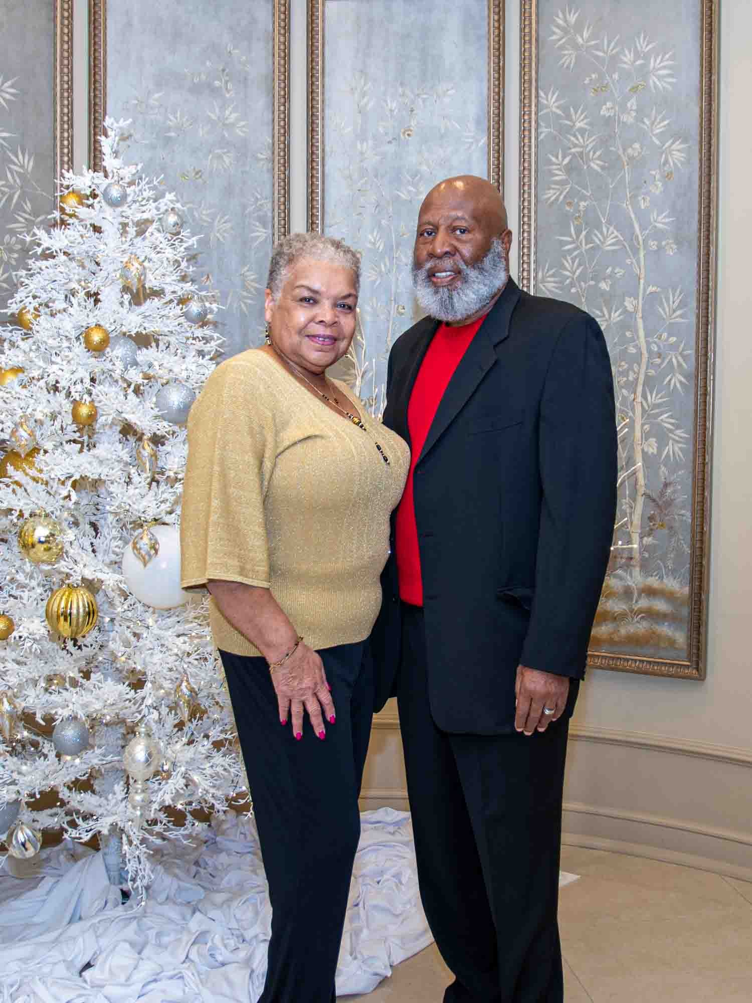 An older couple standing in front of a decorated white Christmas tree with gold and silver ornaments, set against a patterned wall panel.