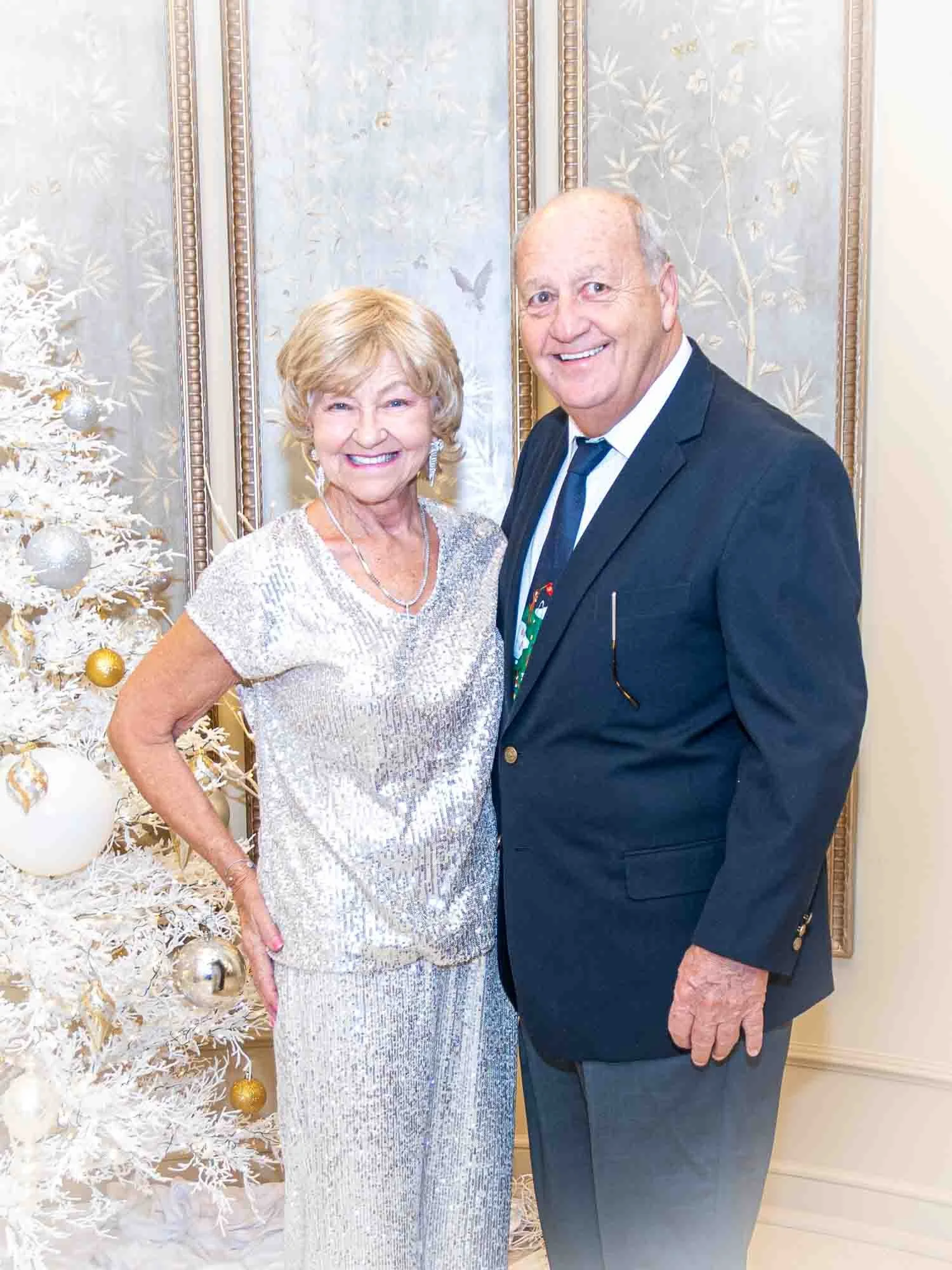 Elderly couple smiling, with a white decorated Christmas tree in the background.