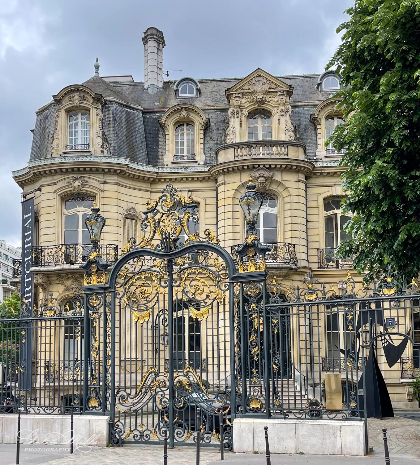 A historic, ornate building with a decorative black and gold wrought iron gate, featuring intricate designs. Two lamp posts are positioned by the gate. The building displays classic architectural style with stone facade, arched windows, and elaborate