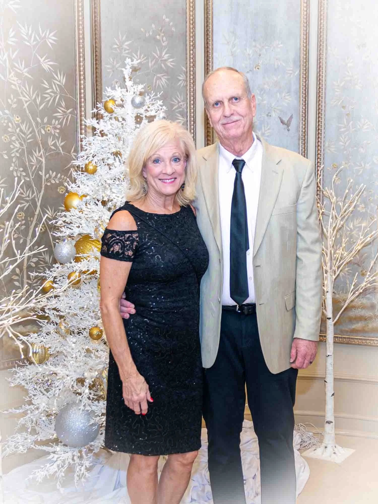 A couple standing in front of a decorative white Christmas tree with golden and silver ornaments, dressed formally, with the woman in a black dress and the man in a light suit and tie.