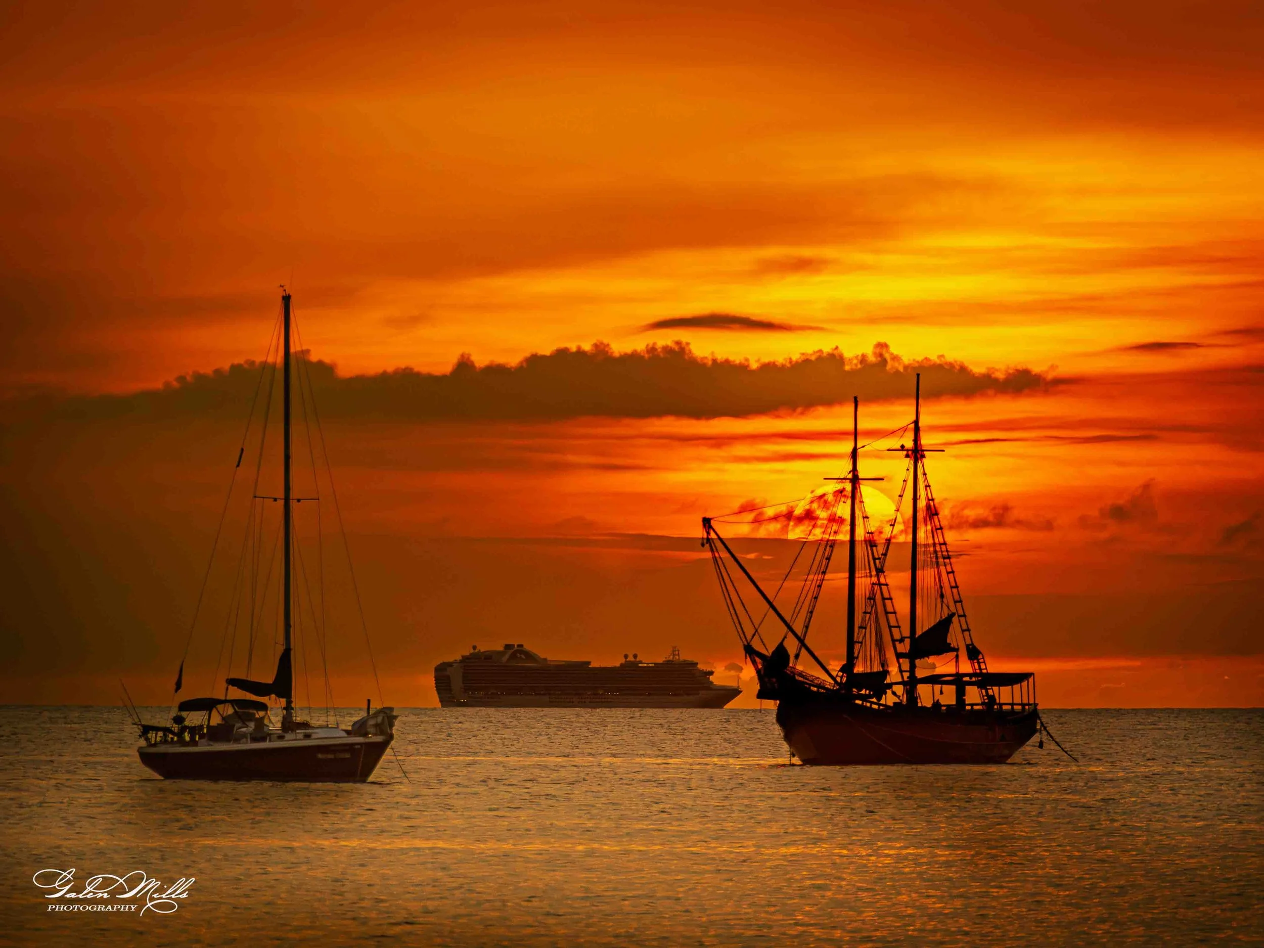 Silhouettes of three boats on the water at sunset, with a cruise ship in the background, and an orange and yellow sky.