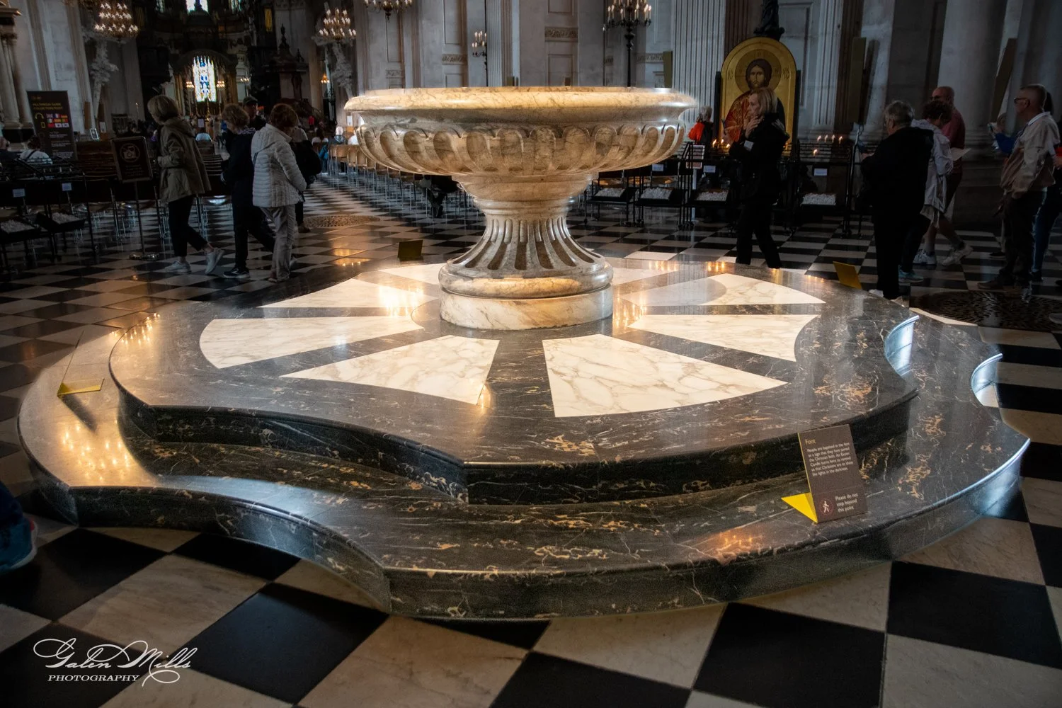 Marble baptismal font in a cathedral interior with checkered floor and visitors.
