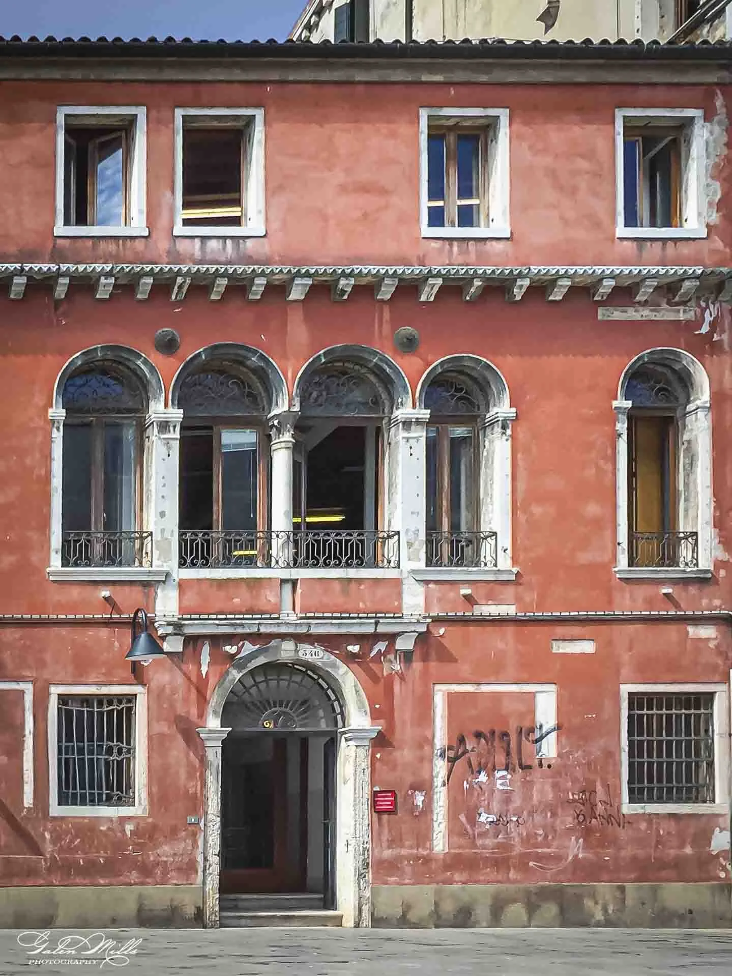 Old, weathered building with a faded red exterior, arched windows and door, balconies with ornate railings, and graffiti at street level.