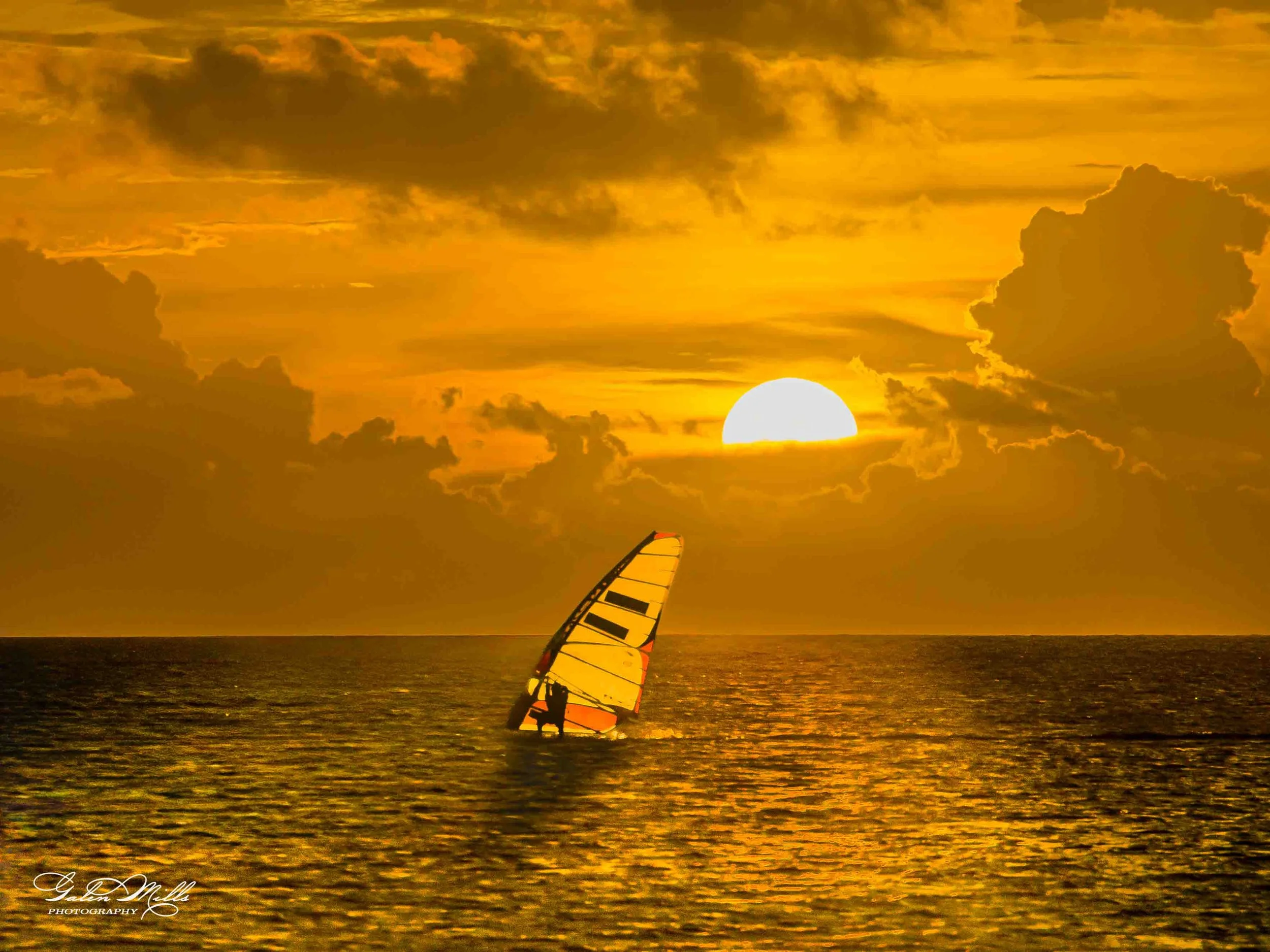 A person windsurfing on the ocean during a golden sunset with partly cloudy sky.