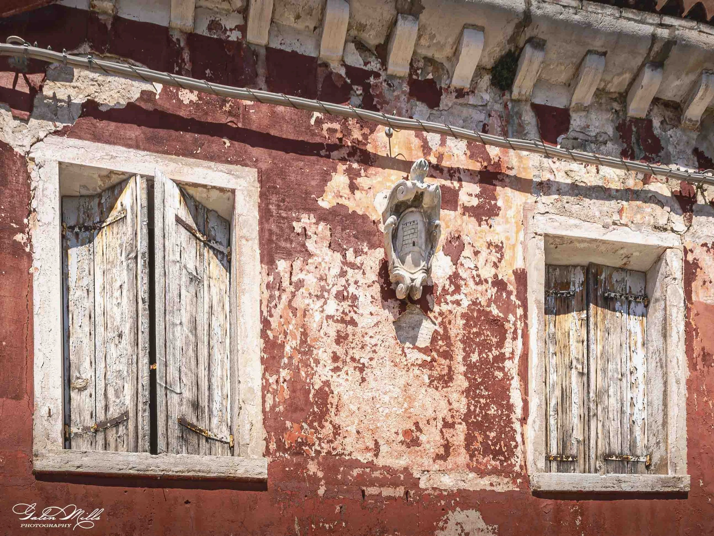 An old, weathered building with peeling red paint, wooden shutters, and a decorative stone face above the window. The building has a row of small, rectangular brick projections along the upper wall and electrical wires running across the facade.