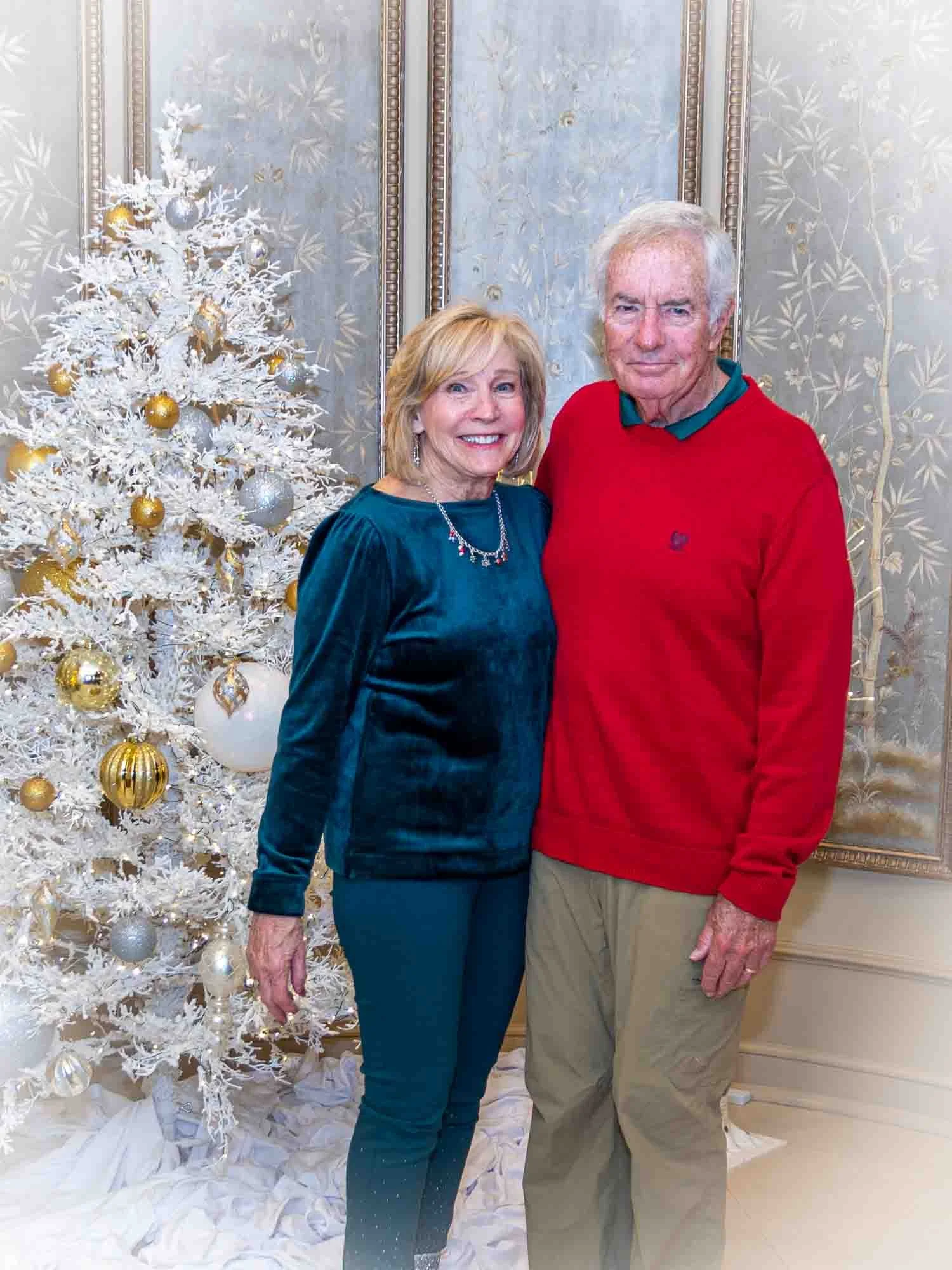 Elderly couple posing in front of a white Christmas tree with gold and silver ornaments, in an elegant room with decorative wallpaper.