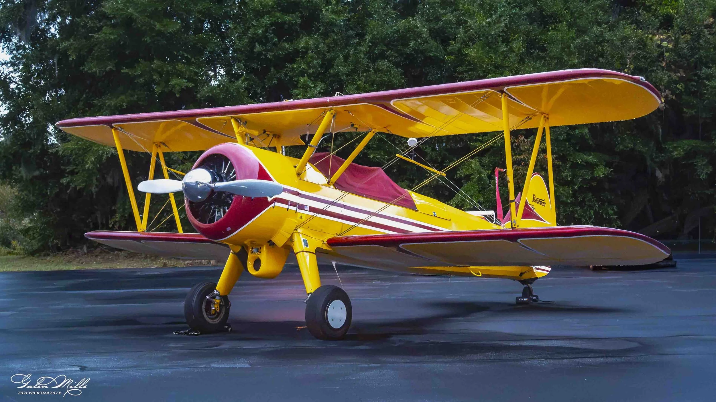 A colorful yellow and red biplane airplane parked on a runway with green trees in the background.