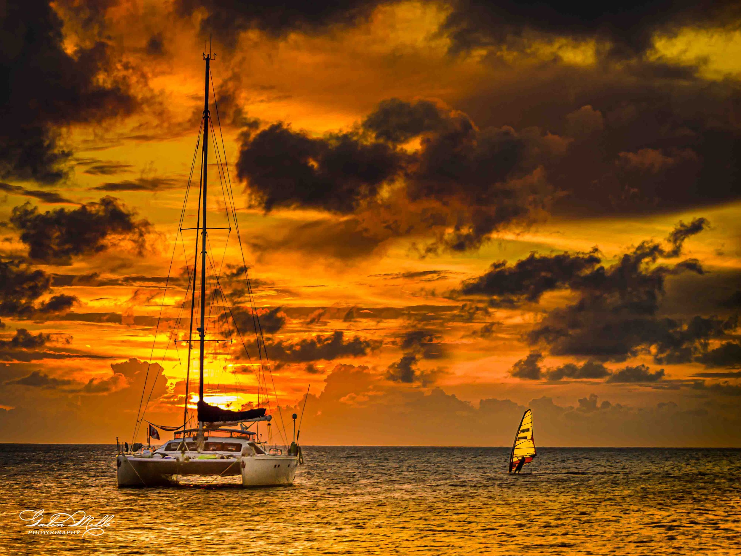 A sailboat and a windsurfer on the ocean during a vibrant sunset with orange and yellow clouds.