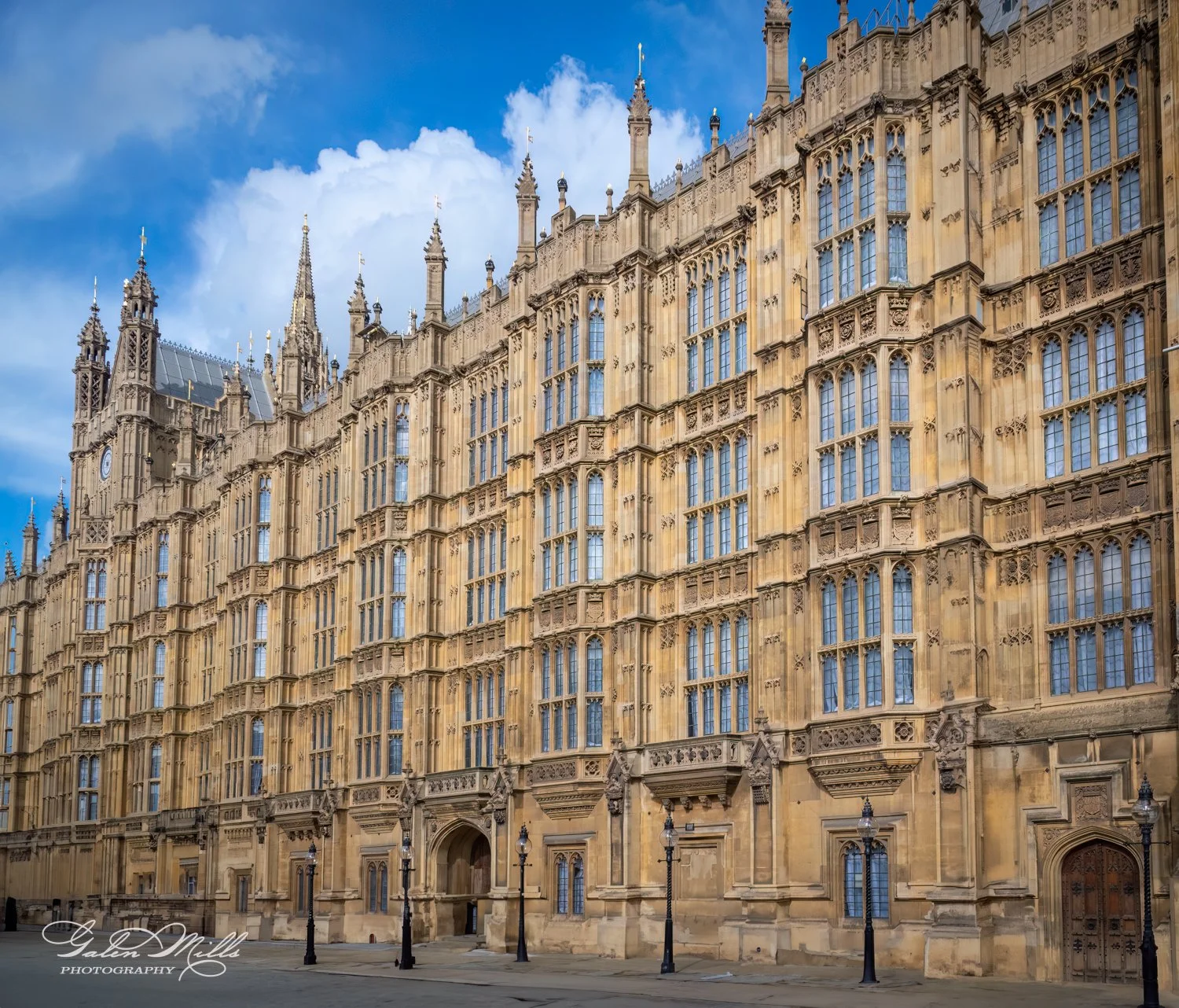 Exterior façade of the Palace of Westminster with Gothic architecture details, large windows, and ornate towers under a blue sky.