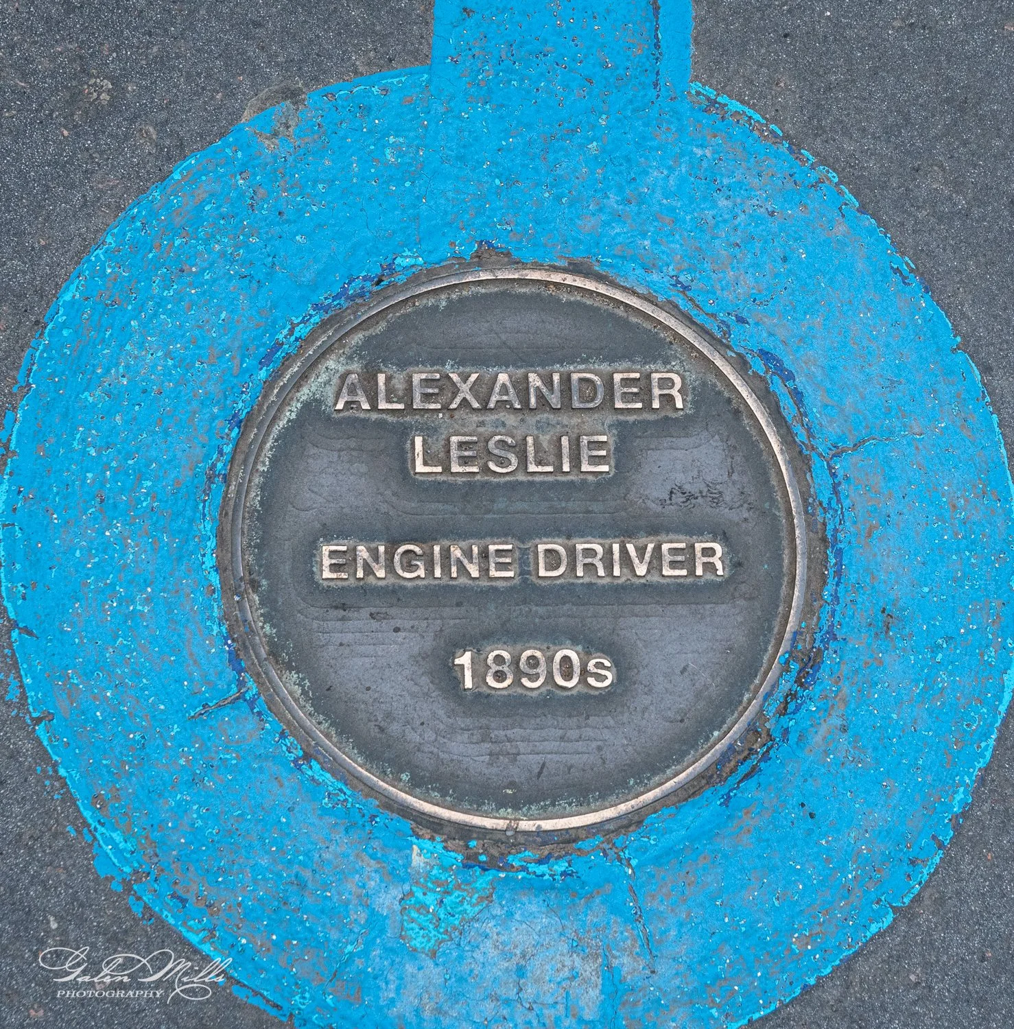 Brass plaque on pavement, "Alexander Leslie, Engine Driver, 1890s," surrounded by blue paint.