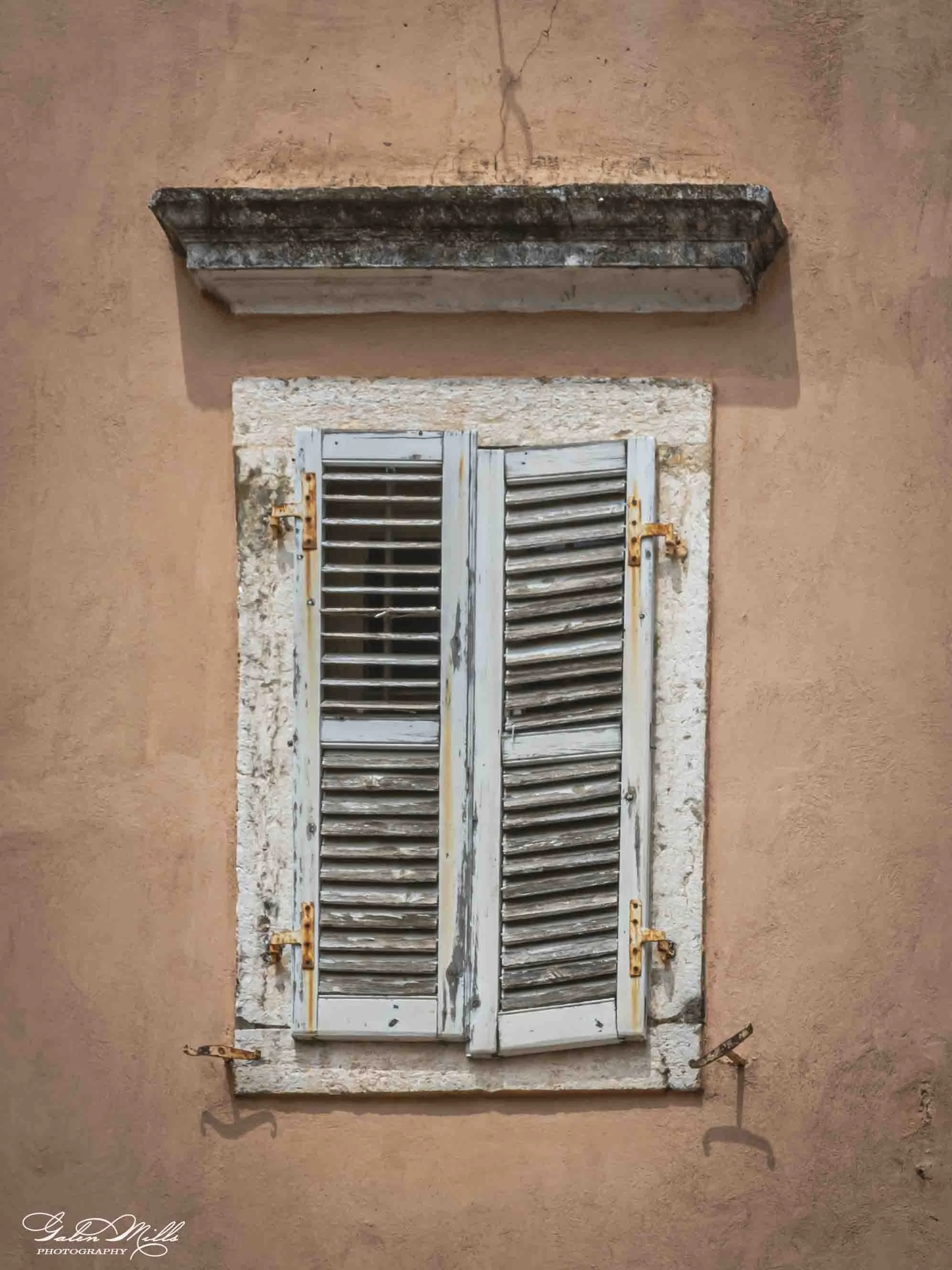 Old white wooden window shutters with louvered slats mounted on a weathered stone frame on a pink adobe wall.