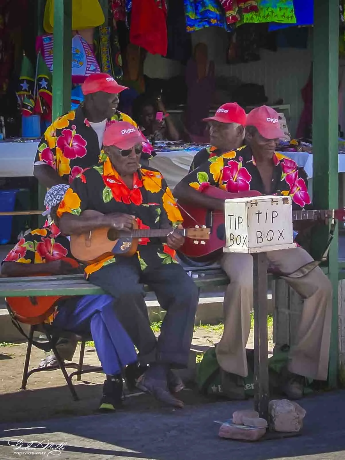 Three men wearing bright floral shirts and red caps with 'Digicel' logo, sitting outdoors on a bench. One man is playing a guitar, while another is sitting beside him. A sign on the table in front reads 'TIP BOX BOX'. There are colorful clothing item