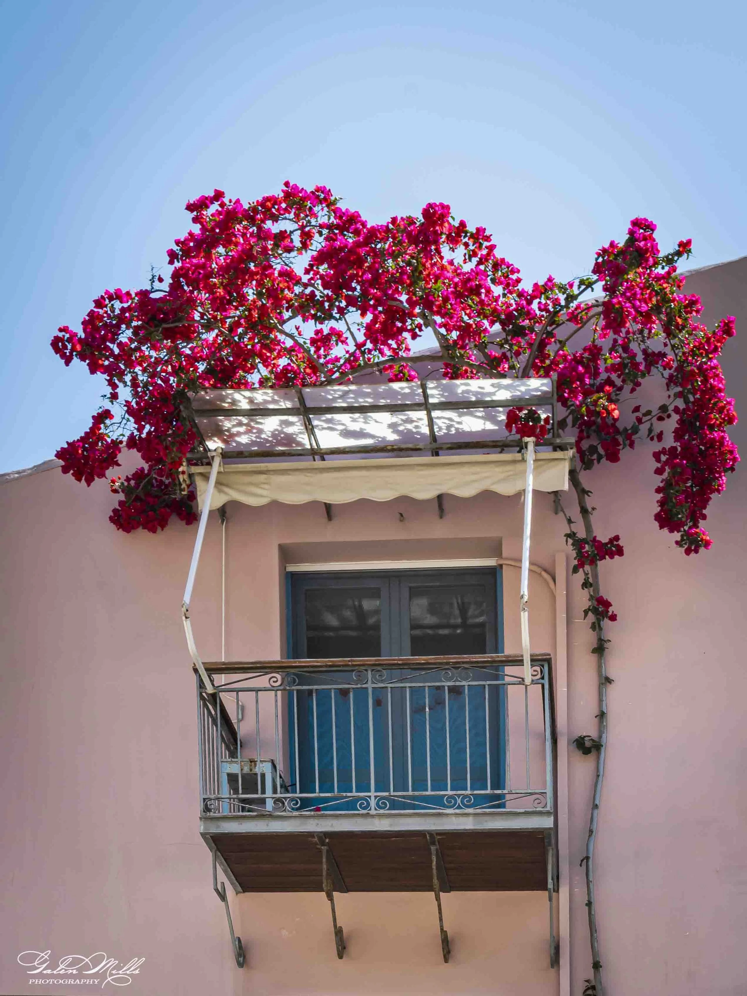 A pink building with a small balcony decorated with blooming pink bougainvillea flowers. The balcony has a blue glass door and a metal railing, with a beige awning above.