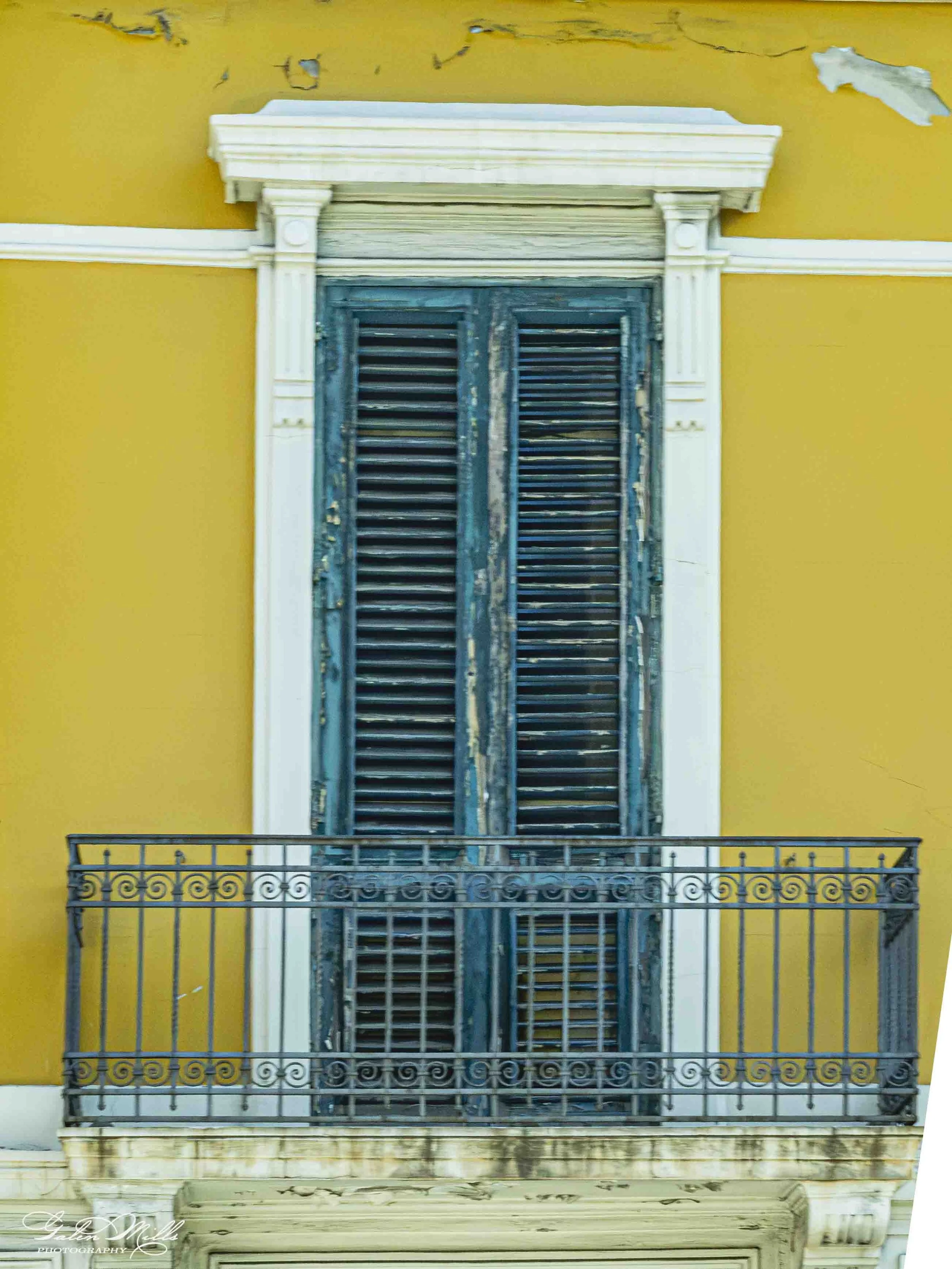 A yellow building facade with an old, weathered blue shuttered window and a small balcony railing below.