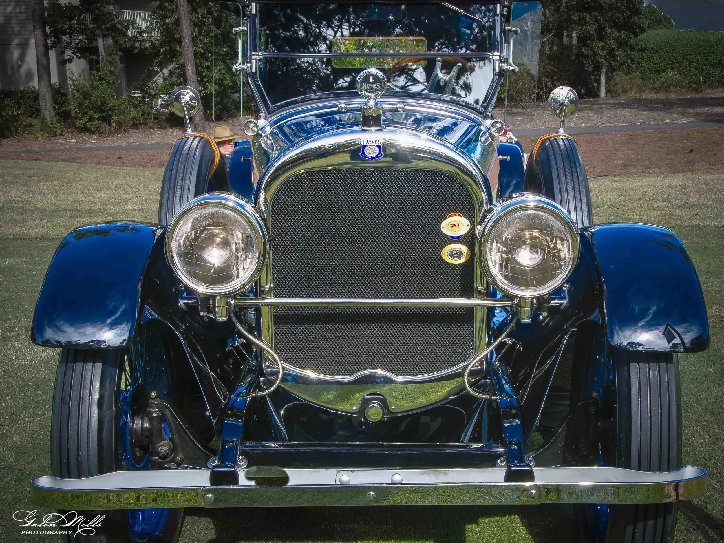 Front view of a vintage black and chrome car with a shiny grill, round headlights, and a Haynes badge, parked outdoors on grass with trees and bushes in the background.