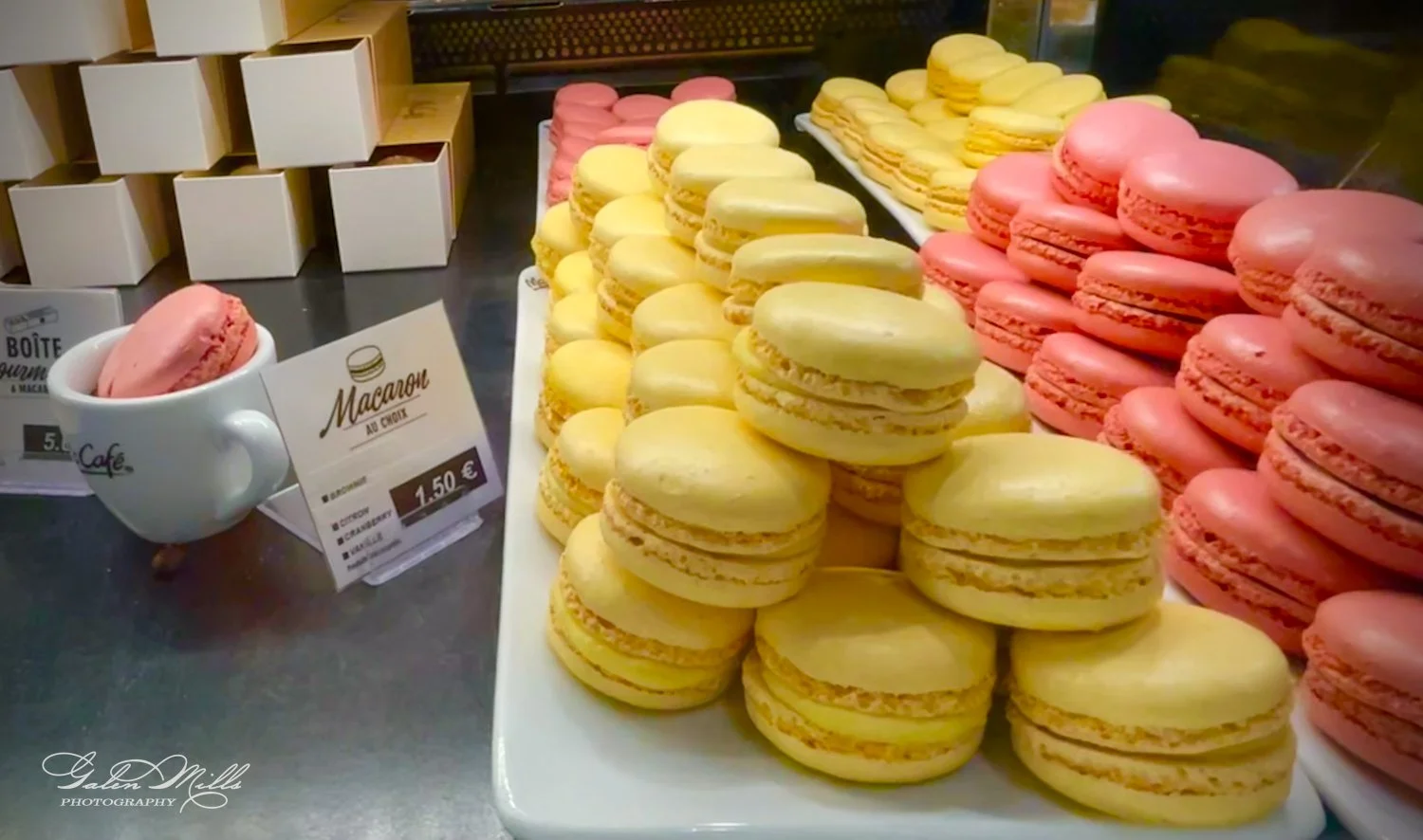 Display of yellow and pink macarons with price signs in a bakery setting.
