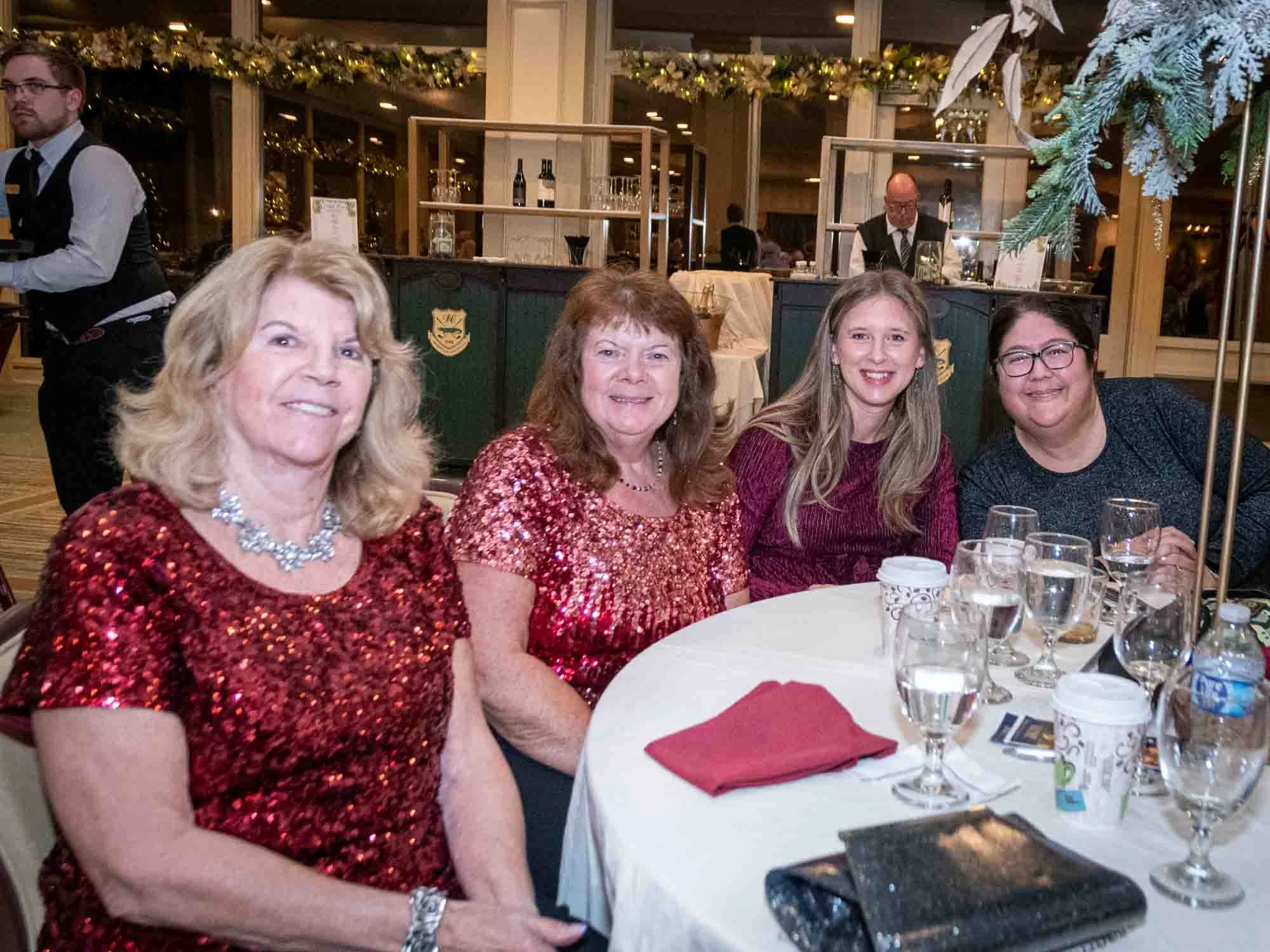 Four women sitting at a table in a festive setting, wearing sparkly tops and smiling. The table is adorned with glasses, cups, and decorations. A waiter is in the background.